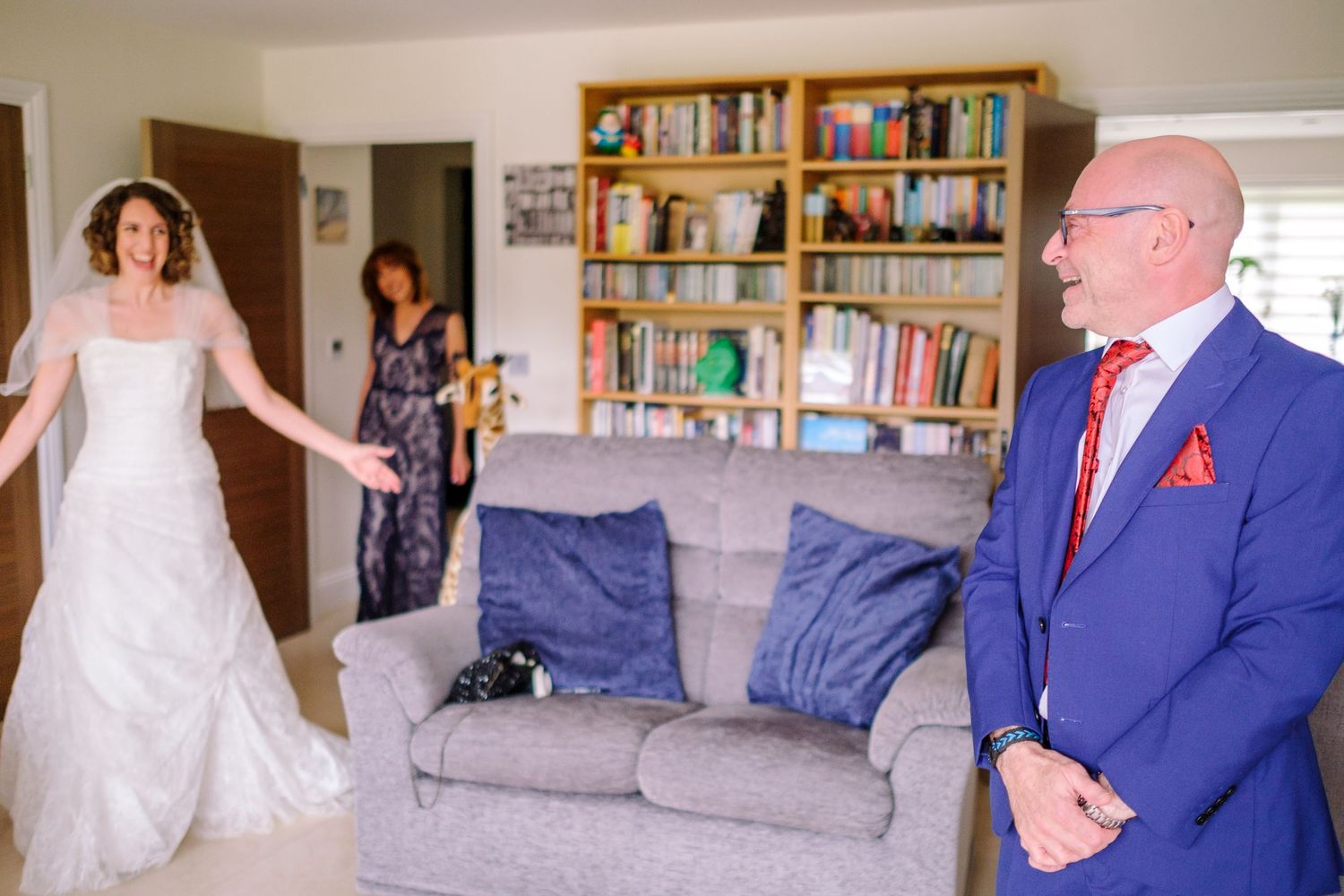 Wedding party members gathered in a living room with bookshelves and blue couch for pre-ceremony preparations.