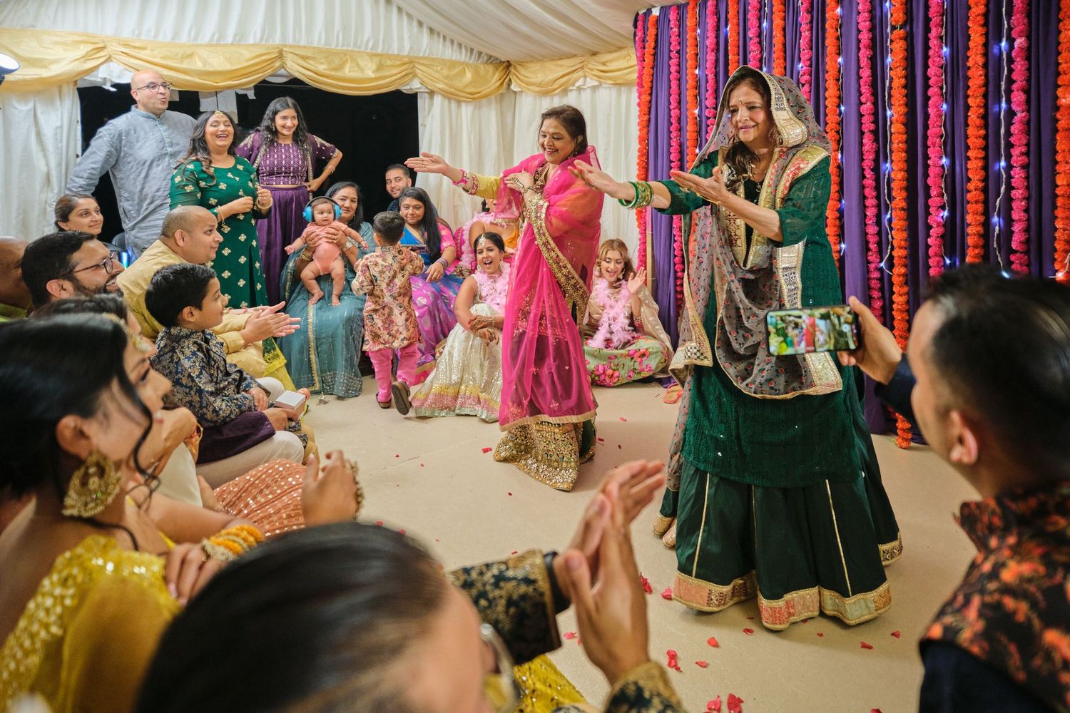 Guests celebrate at a colorful Indian wedding ceremony with vibrant pink and purple decorations in a tent setting.