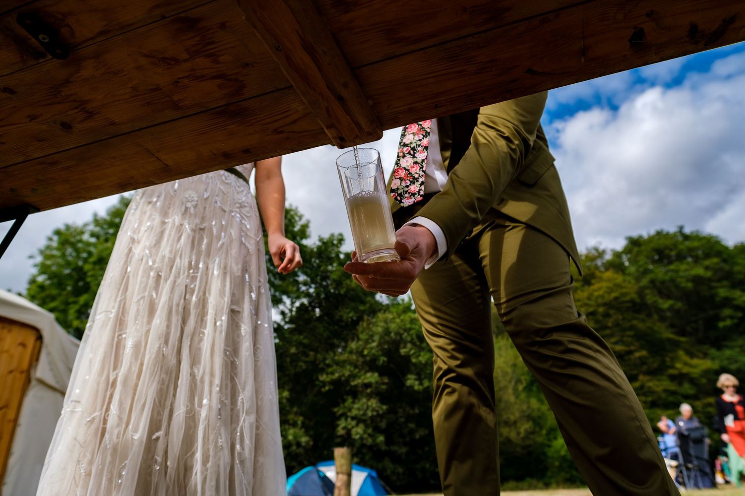 A couple toasts with drinks at an outdoor wedding celebration with blue skies and trees in the background.
