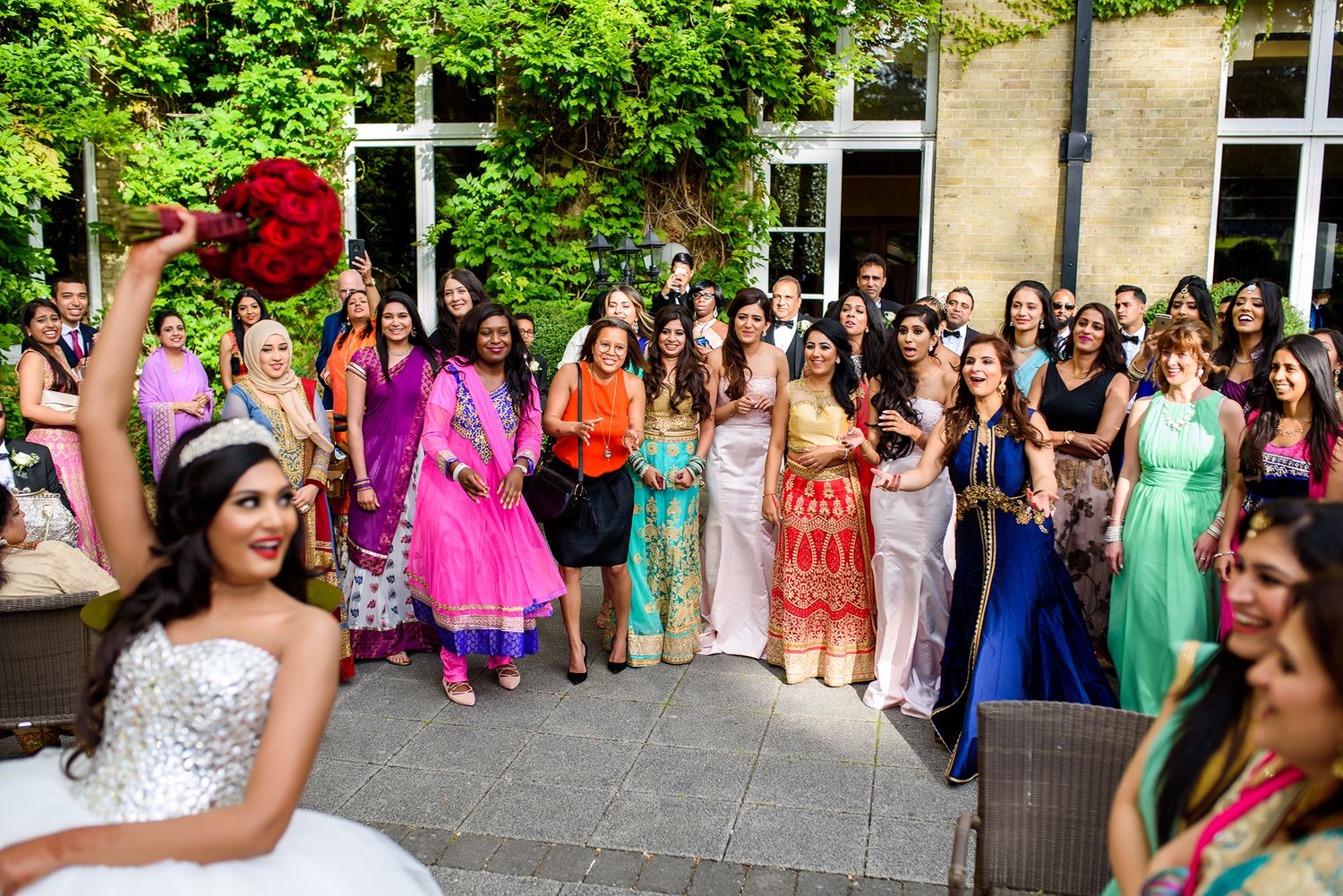 Large wedding group photo with colorful dresses outdoors by a stone building.