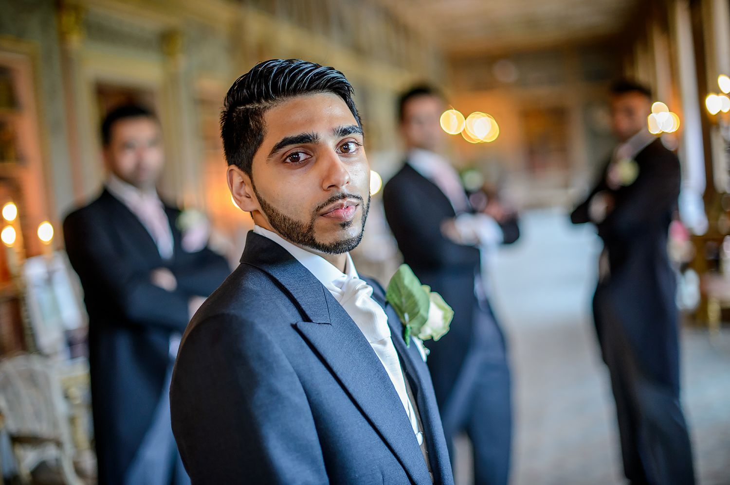 Groom in grey suit with boutonniere stands in elegant hallway.