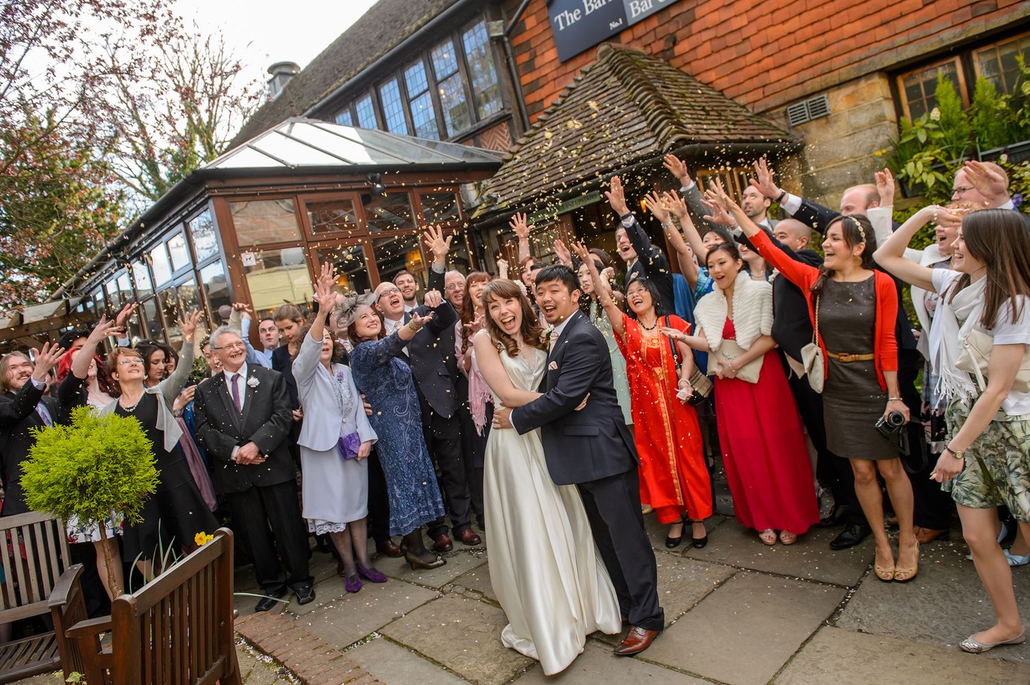 Gathered wedding guests celebrate outside a brick building with red roof tiles.