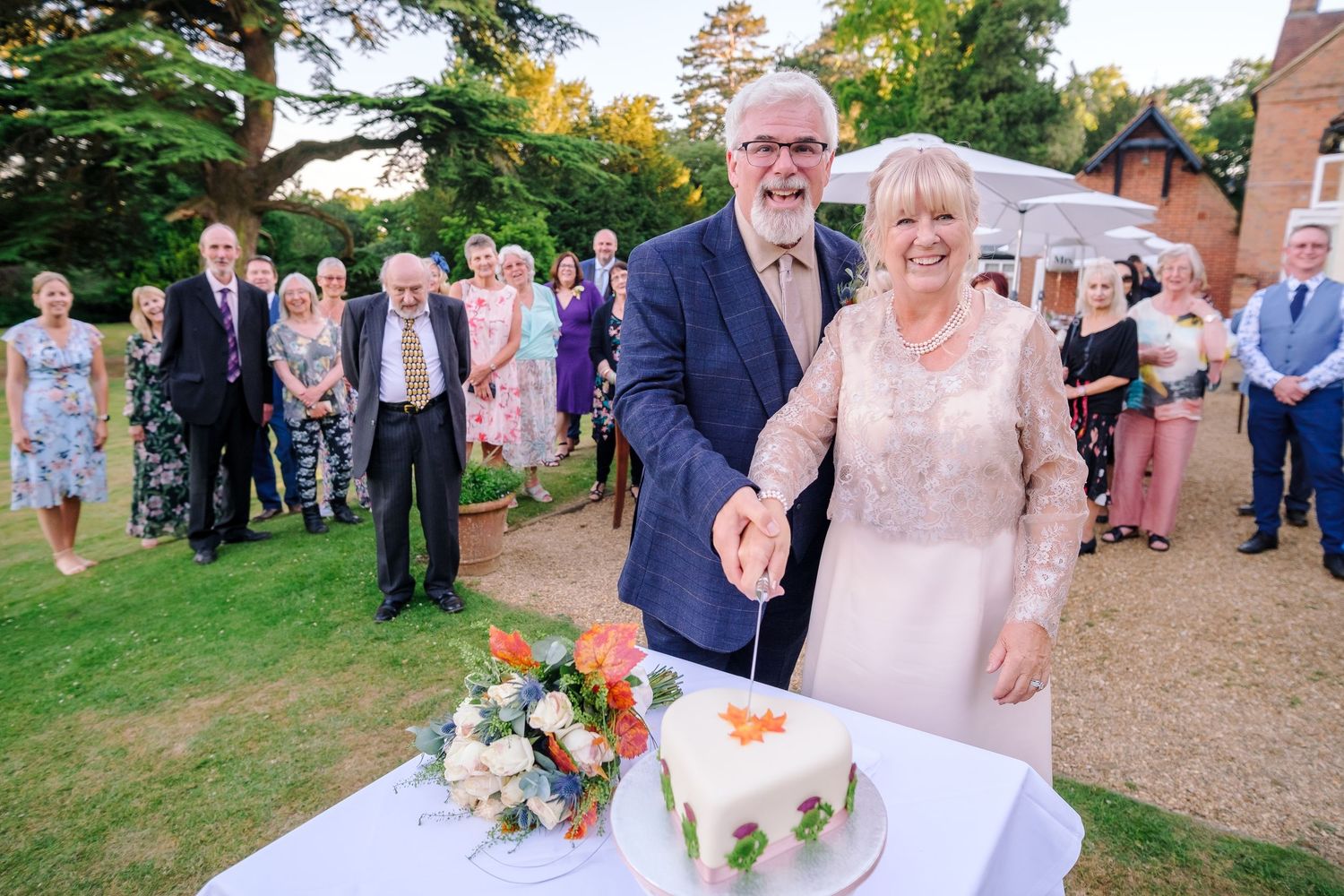 A wedding celebration scene with guests gathered around a decorated cake table outdoors.