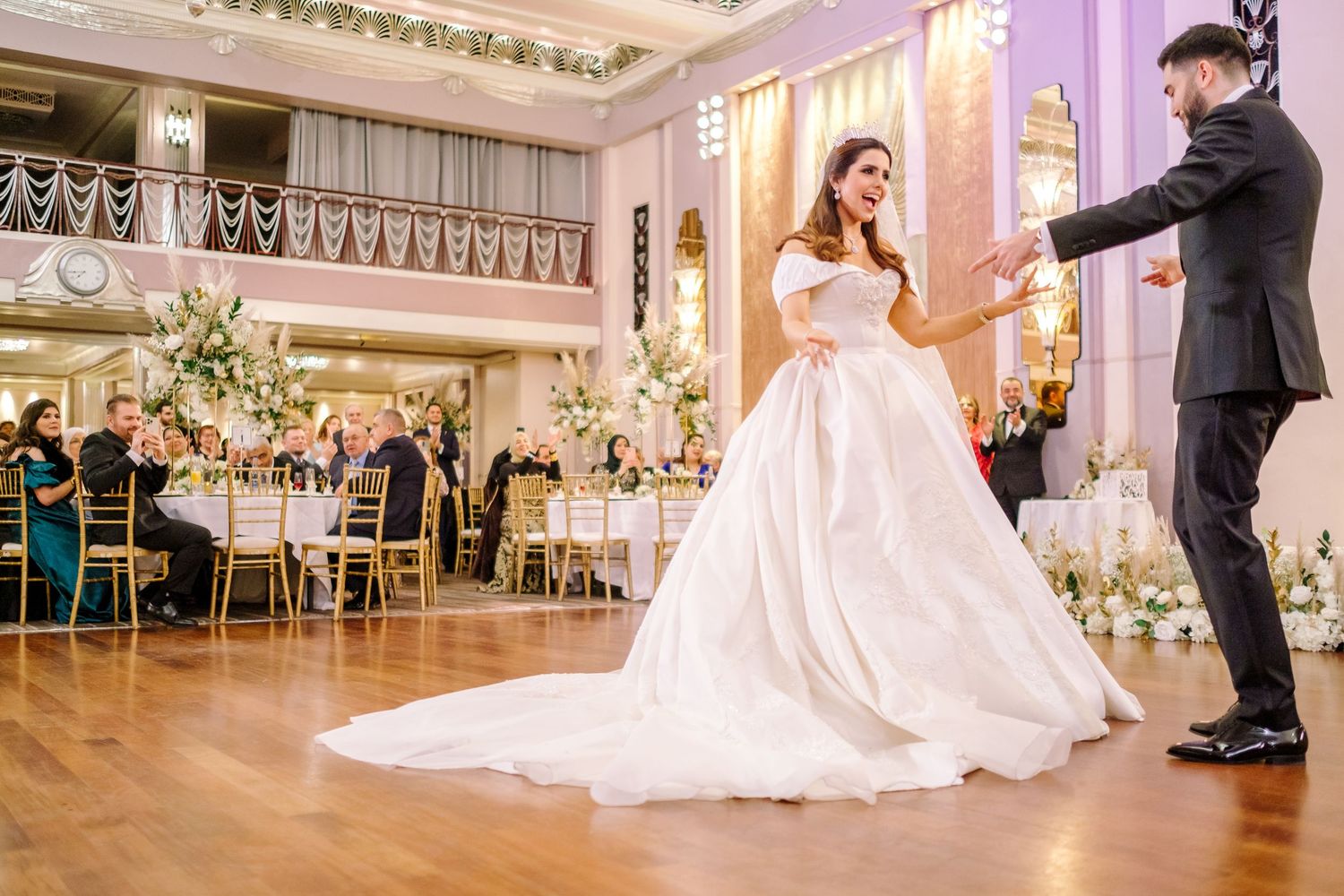 A couple shares their first dance in an elegant ballroom decorated with floral arrangements and twinkling lights.