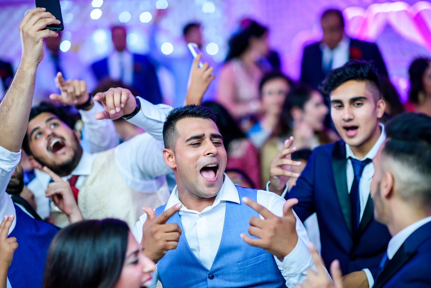 Energetic wedding party crowd cheering and celebrating under vibrant purple lights.