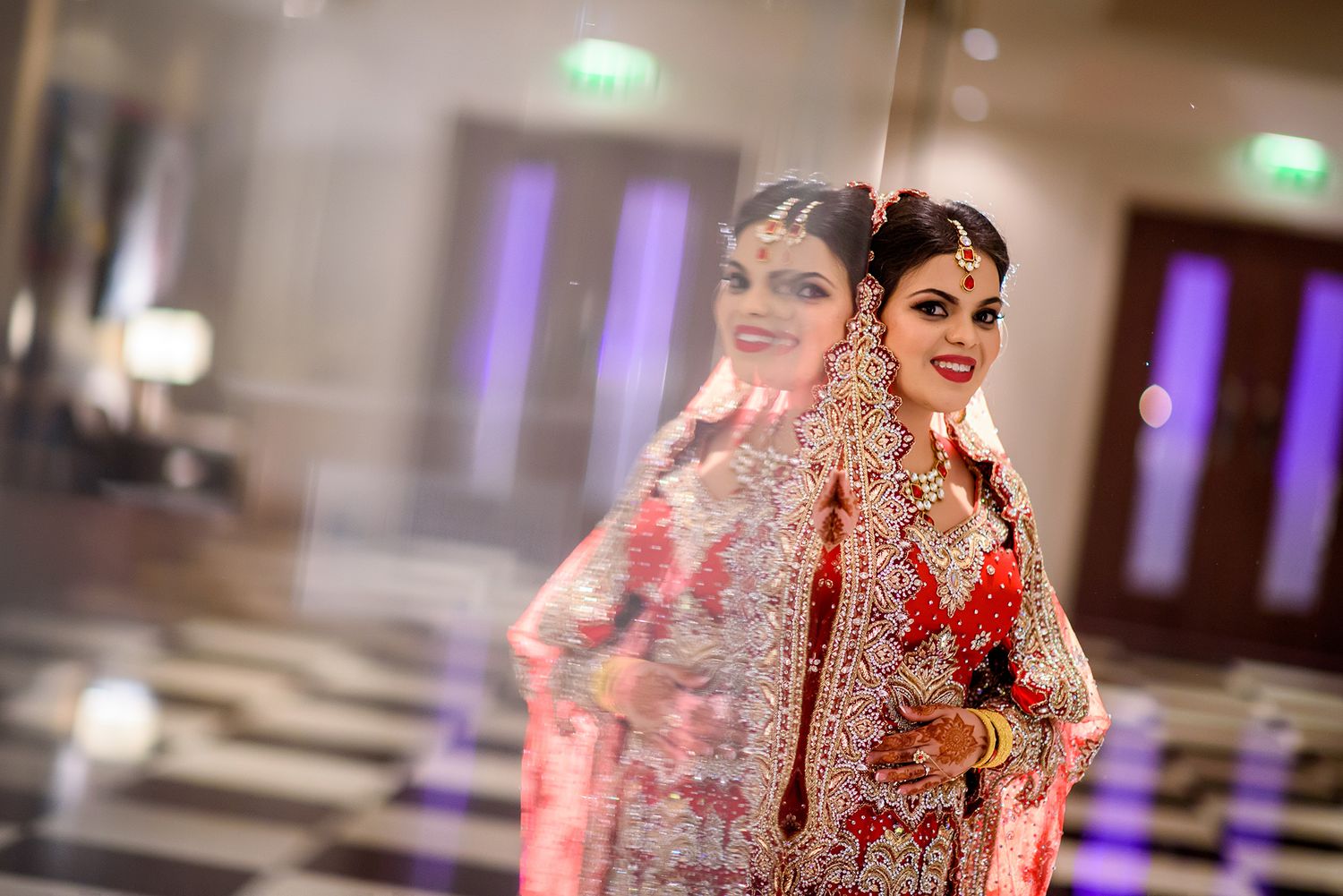 Traditional South Asian bride in elegant red and gold wedding attire poses gracefully in an indoor venue.