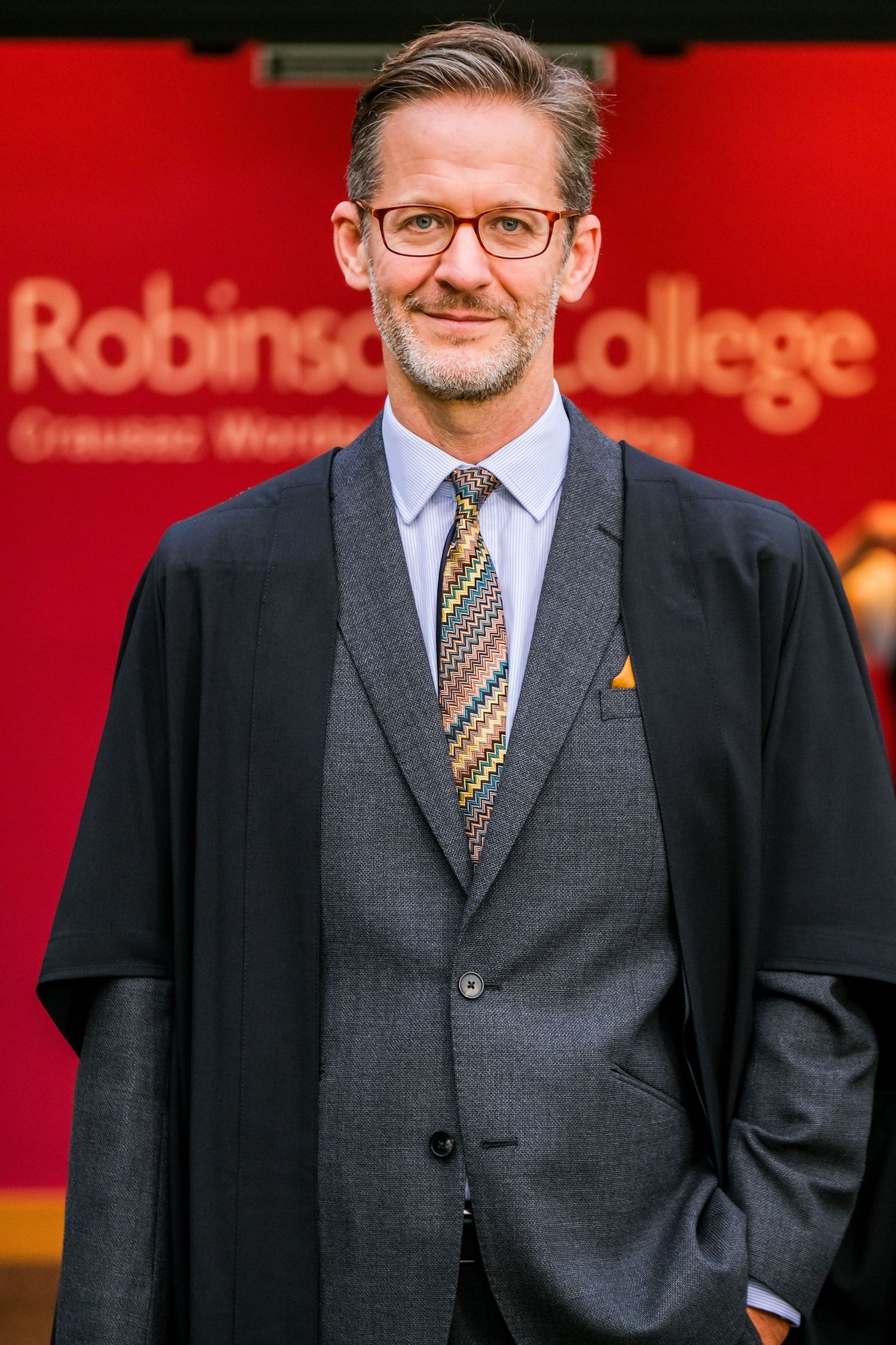 A person in a dark gray suit and tie stands in front of a red background with a warm expression.
