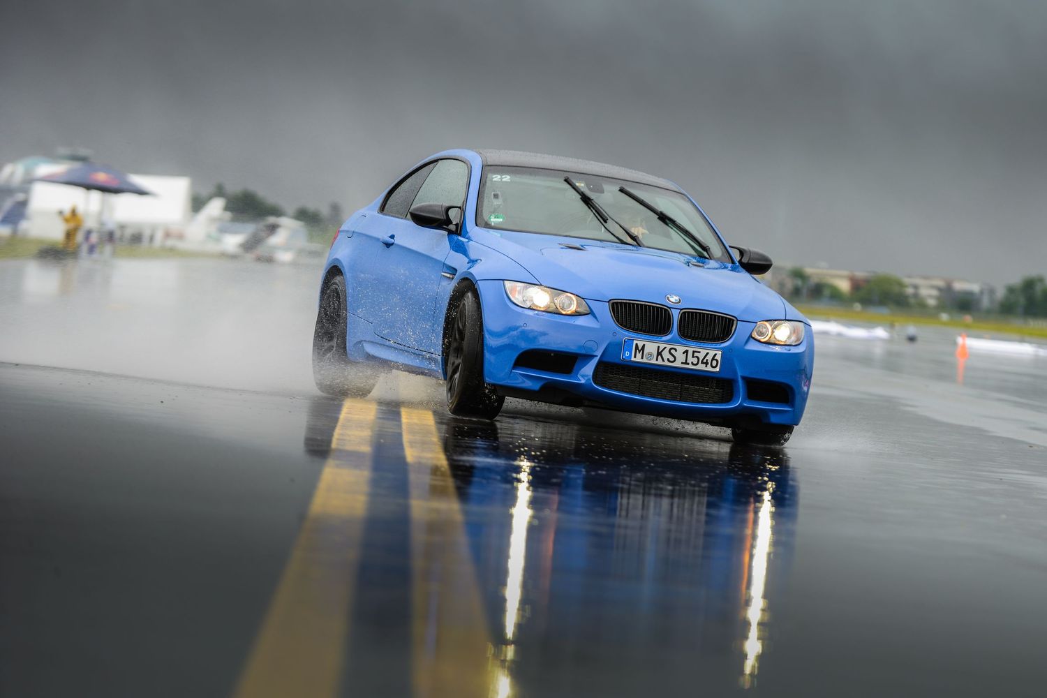 Blue BMW sports car driving on wet pavement with reflection in rain puddles.