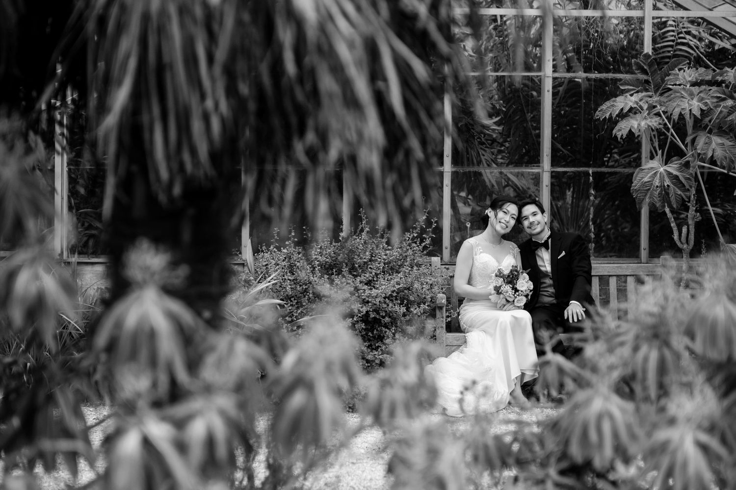 Black and white wedding photos taken through tropical foliage in garden setting.