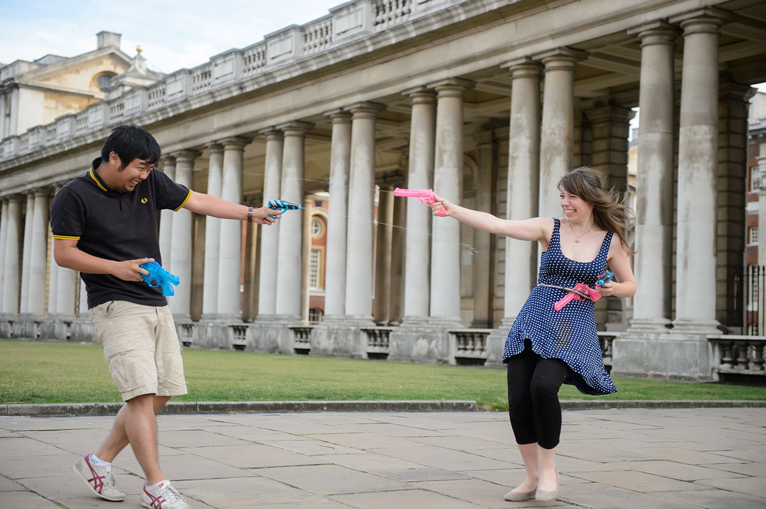 Two people playfully pose with arms outstretched in front of a historic colonnade.
