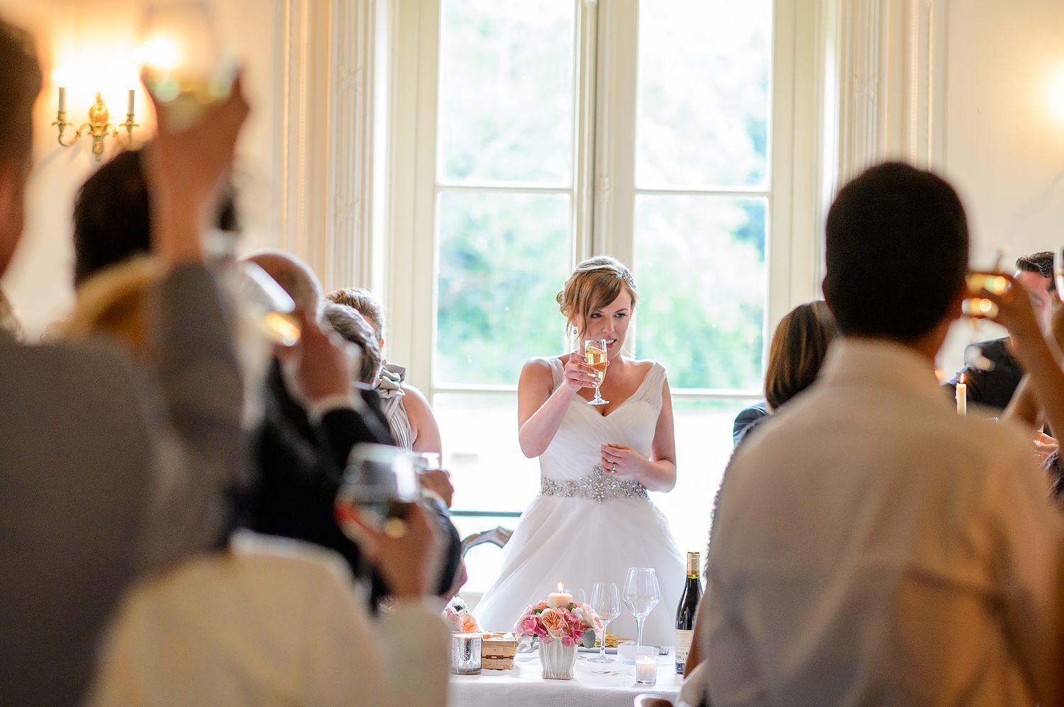 Wedding reception guests mingle in a warmly lit room with tall windows.