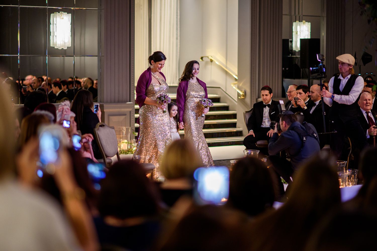 Guests capture photos of a wedding celebration on their phones in an elegant venue.