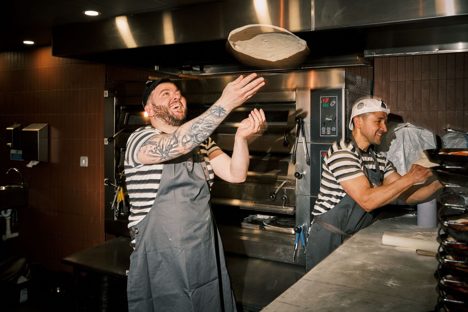 Two chefs in striped shirts and gray aprons toss pizza dough in a restaurant kitchen.