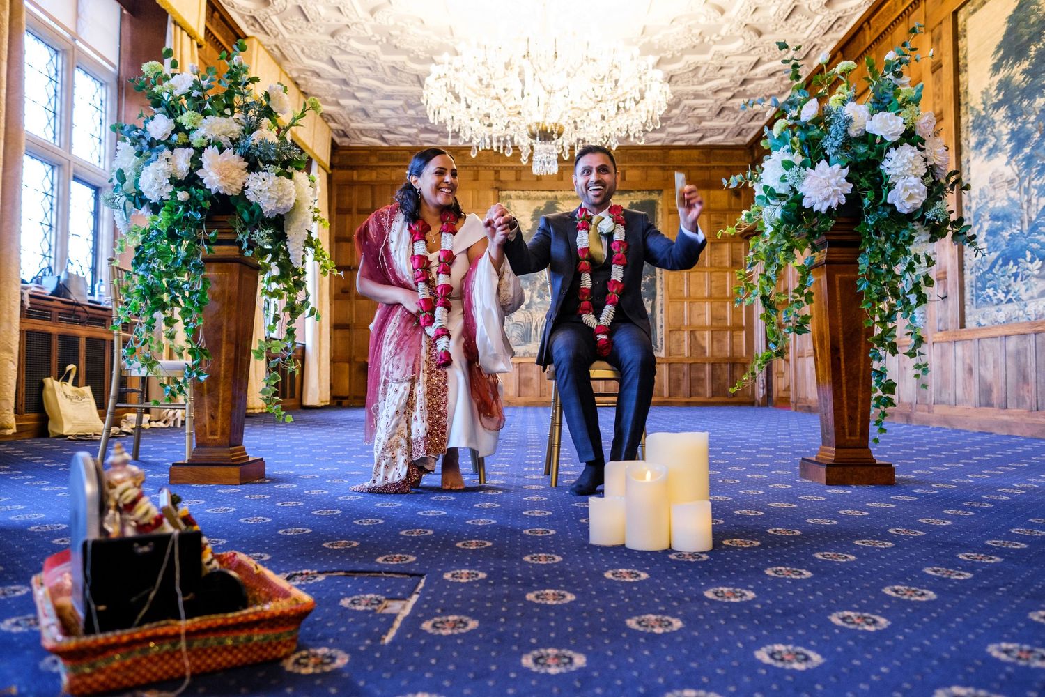 Wedding guests gather in an elegant ballroom decorated with floral arrangements and ornate carpeting.