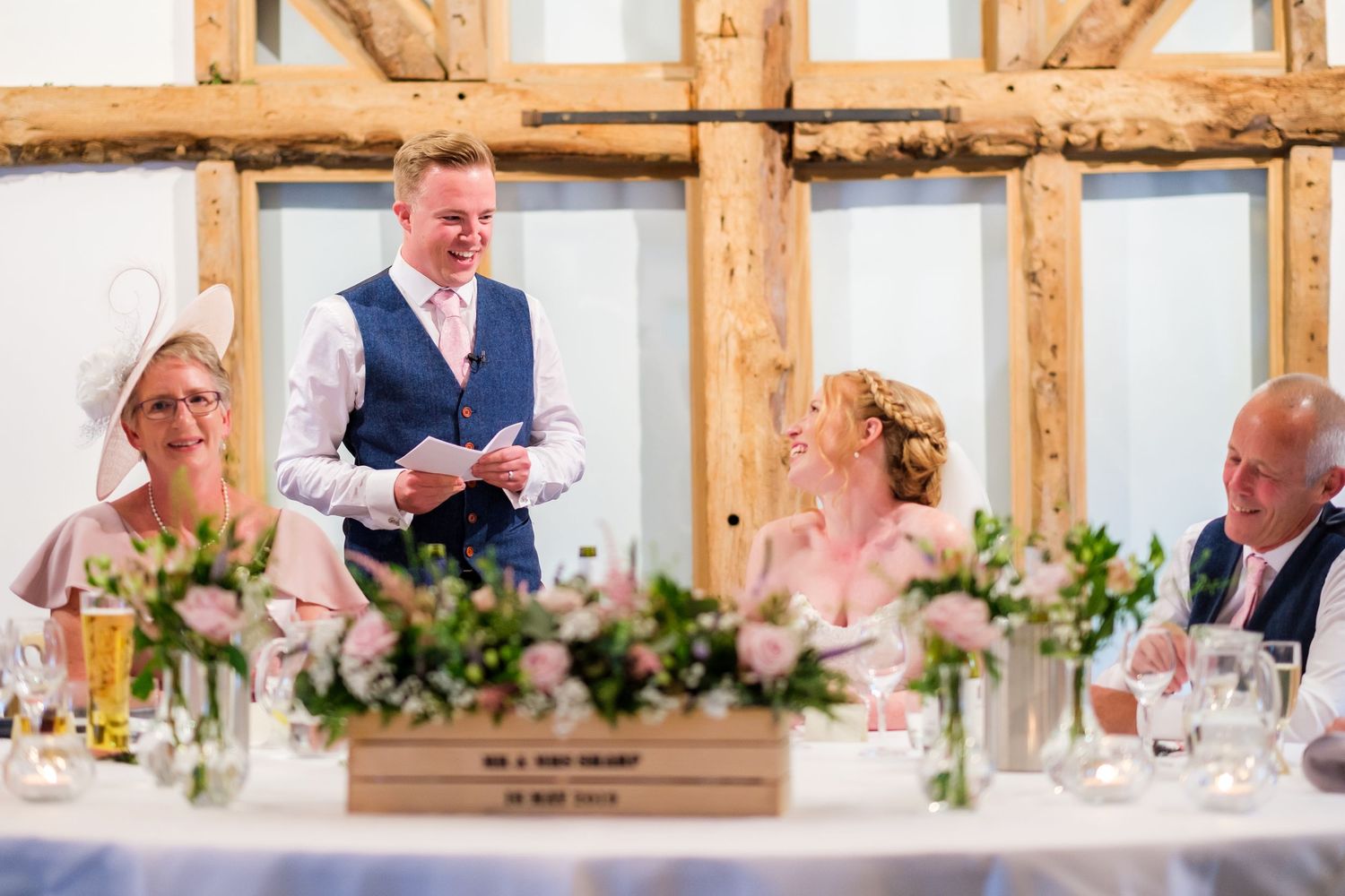 Wedding reception speech in a timber-framed venue with elegant floral centerpieces on dining tables.