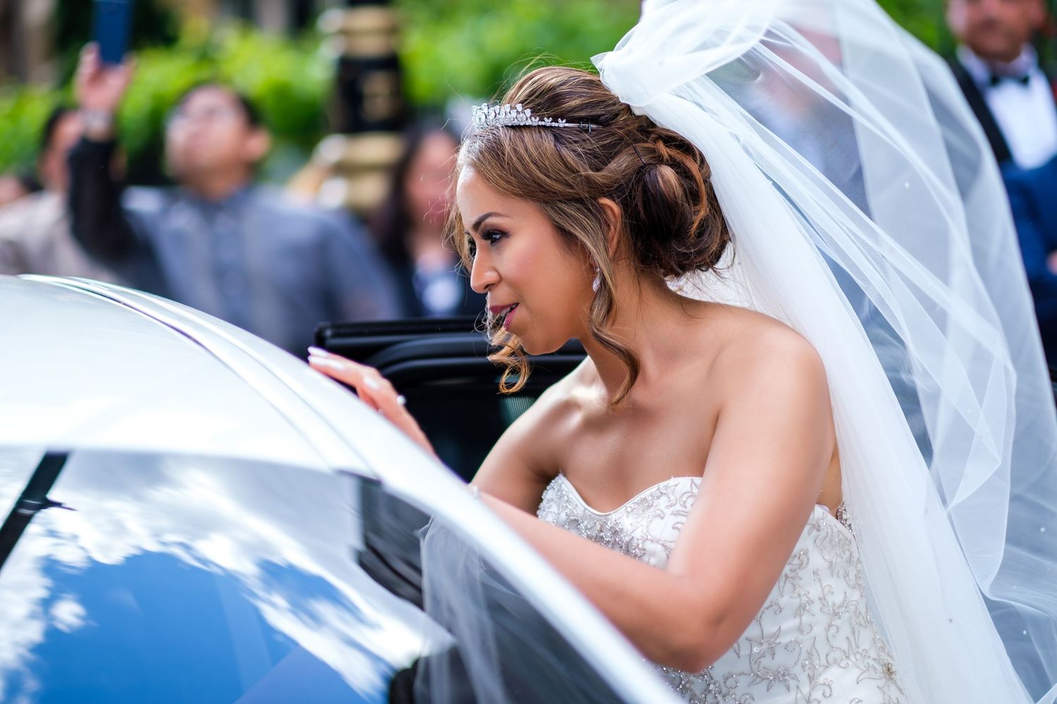 A person in a wedding dress with flowing veil enters a white vehicle.