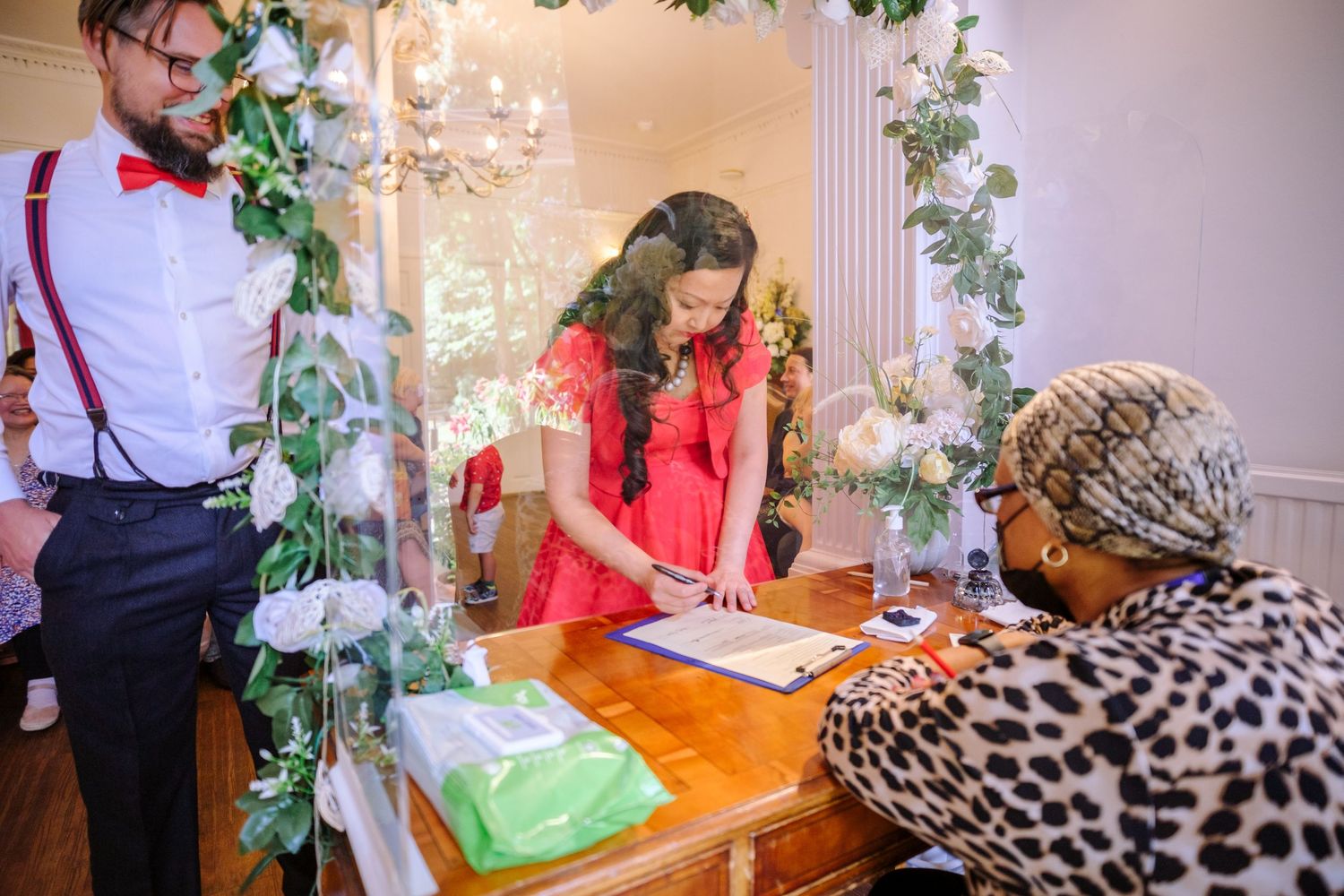A couple signs a document at an elegant wedding venue decorated with greenery and chandeliers.
