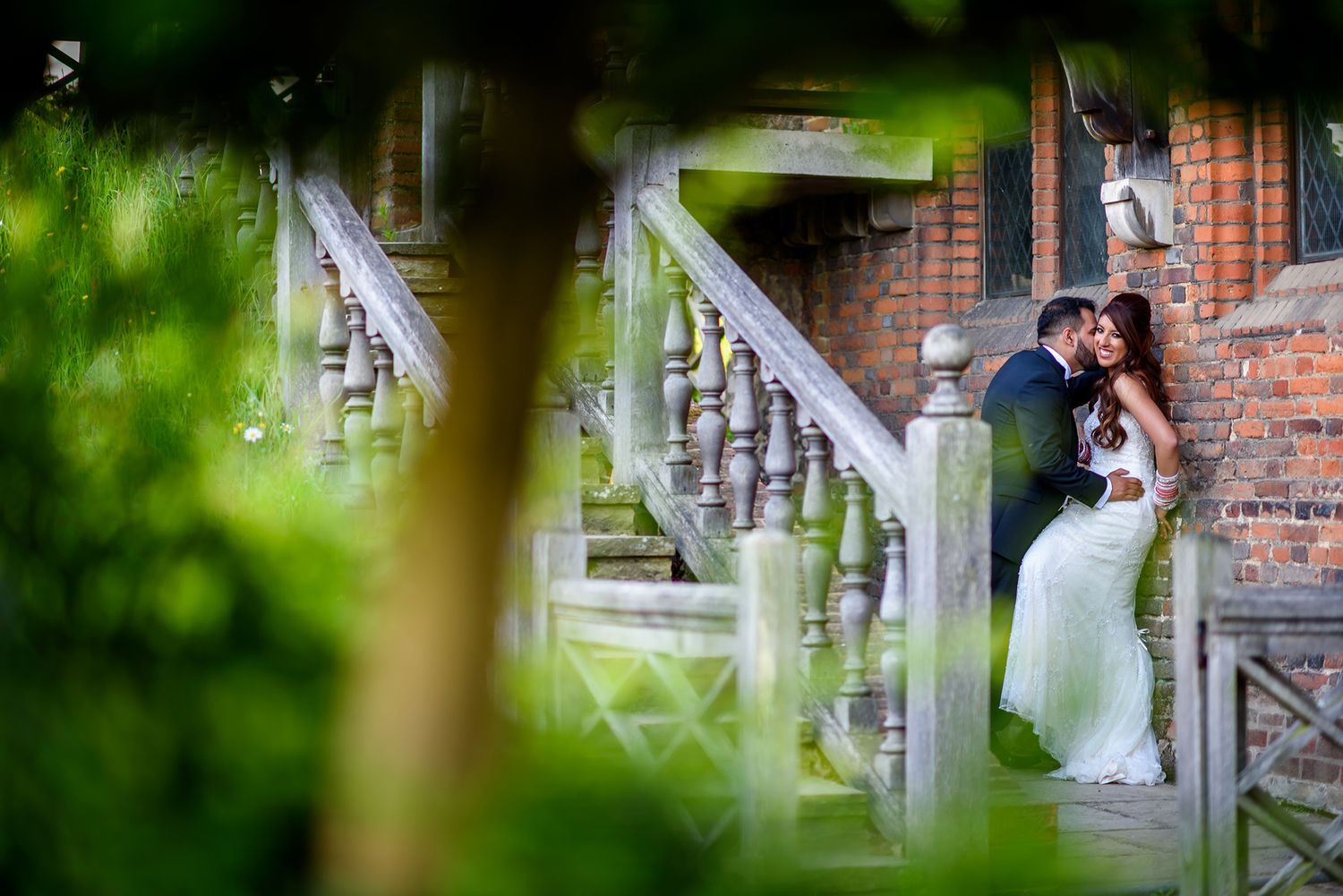 An elegant wedding couple shares an intimate moment against a rustic brick wall backdrop in an outdoor garden setting.