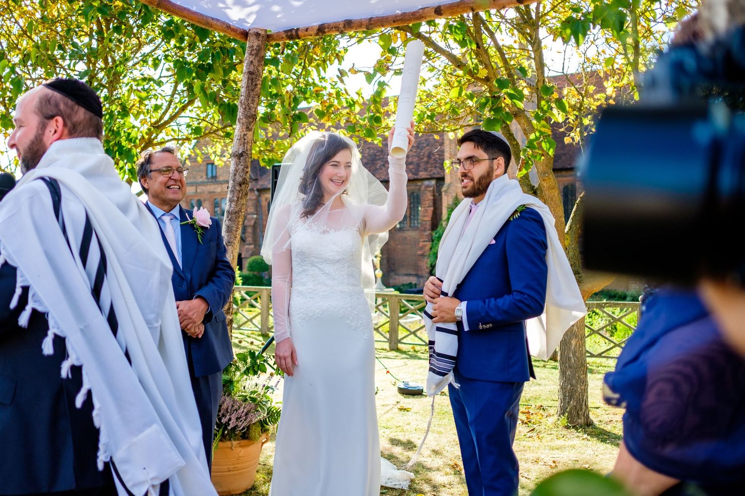Wedding ceremony taking place in sunlit garden with trees and white decor.