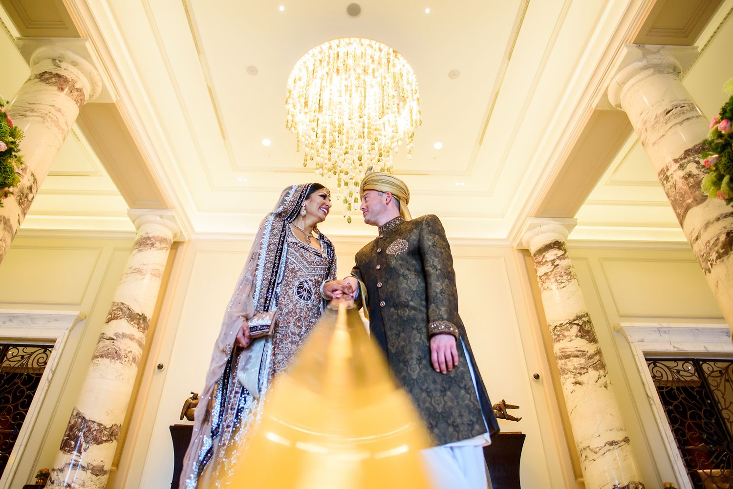 Elegant couple shares a tender moment under an ornate chandelier in a luxurious venue.