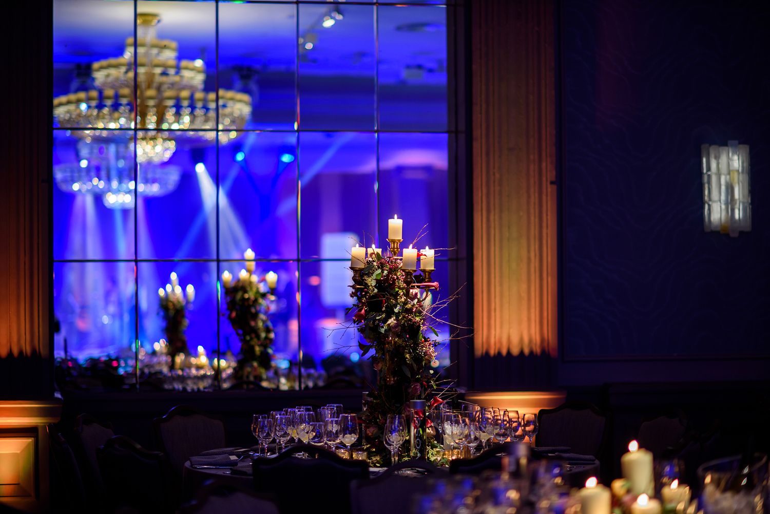 Elegant wedding reception venue with blue uplighting and ornate crystal chandeliers illuminating round dining tables.