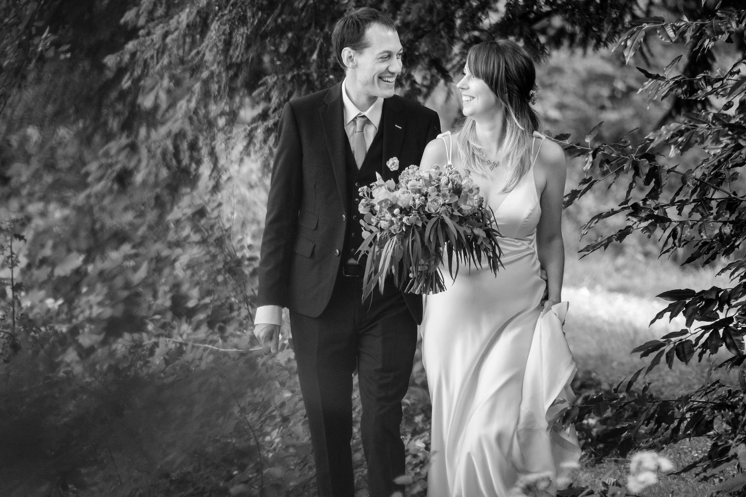Elegant wedding couple walks through garden pathway surrounded by greenery.