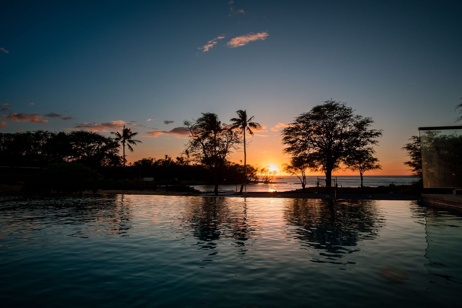 Beautiful tropical sunset reflected in calm infinity pool with palm tree silhouettes against orange sky.