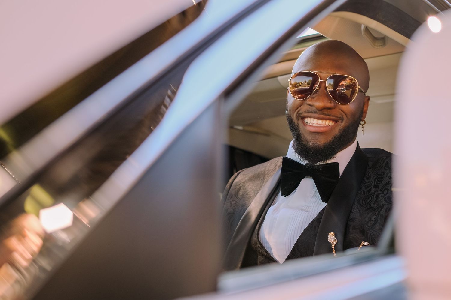 Groom in sunglasses smiles joyfully while seated in a car on his wedding day.