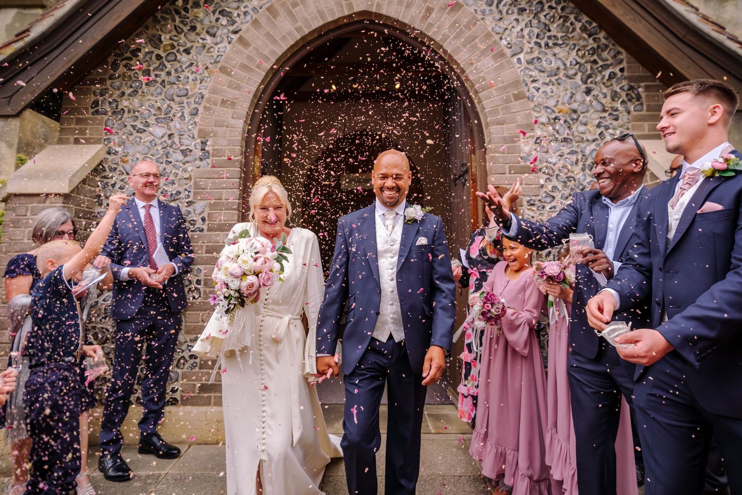 Wedding guests throw confetti outside a stone church archway during a joyous celebration.
