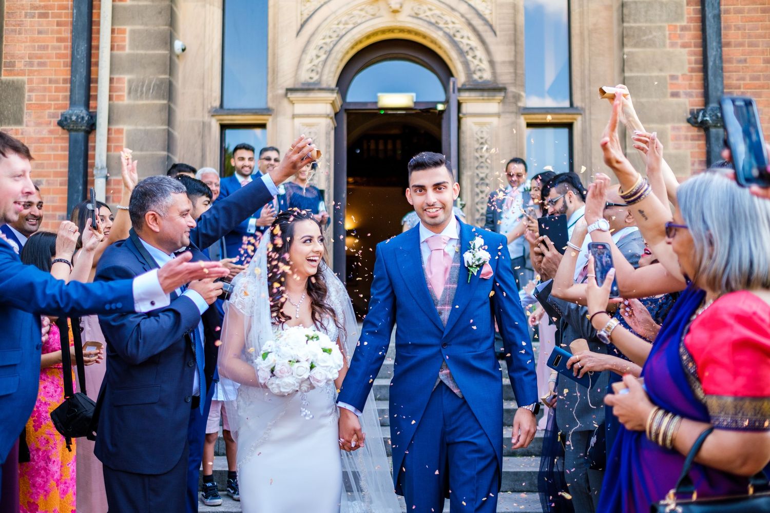 Wedding guests throw confetti as newlyweds exit church while celebrating in bright blue and purple attire.