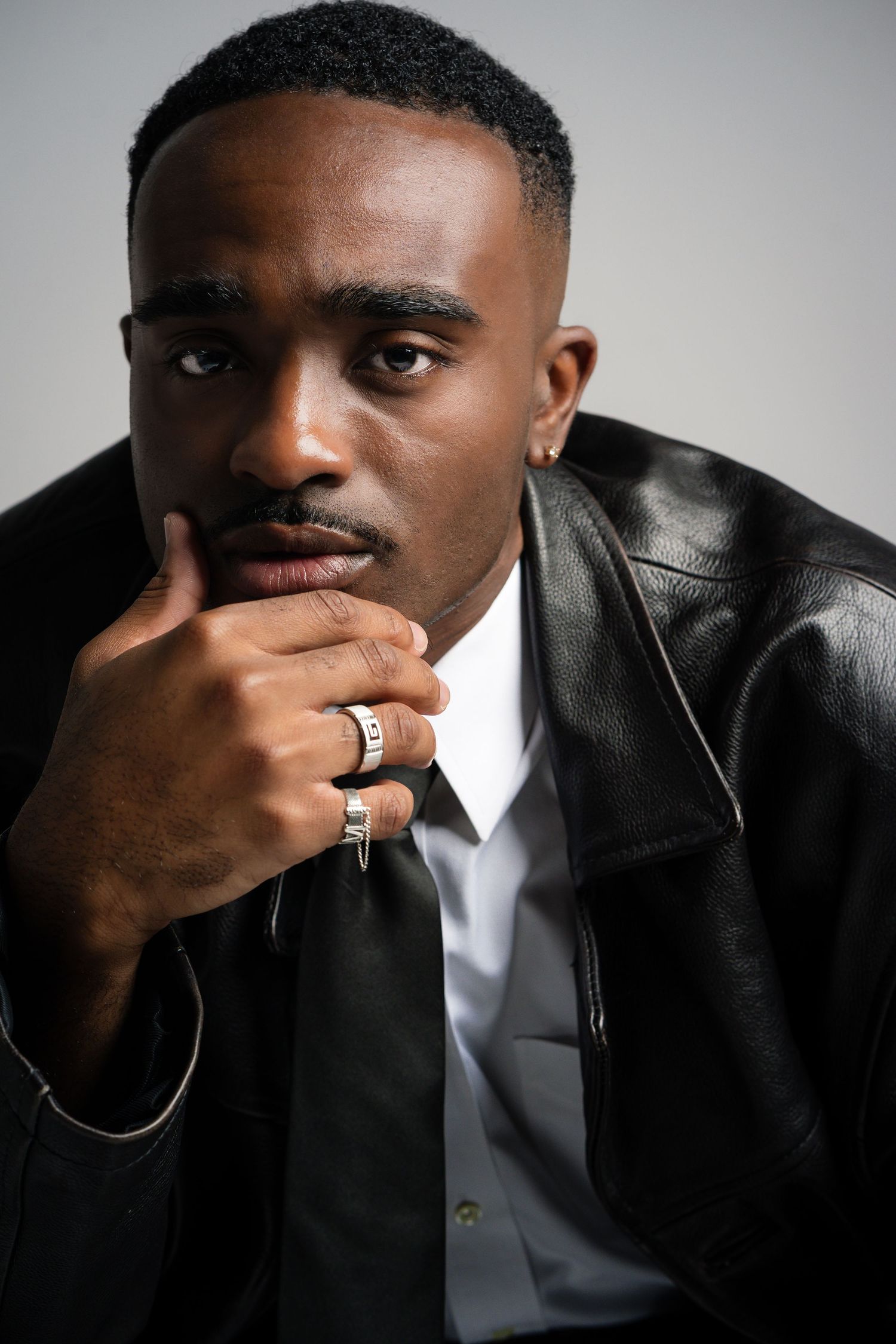 Young Black man in leather jacket and white shirt poses thoughtfully with hand on chin, wearing rings and earring.