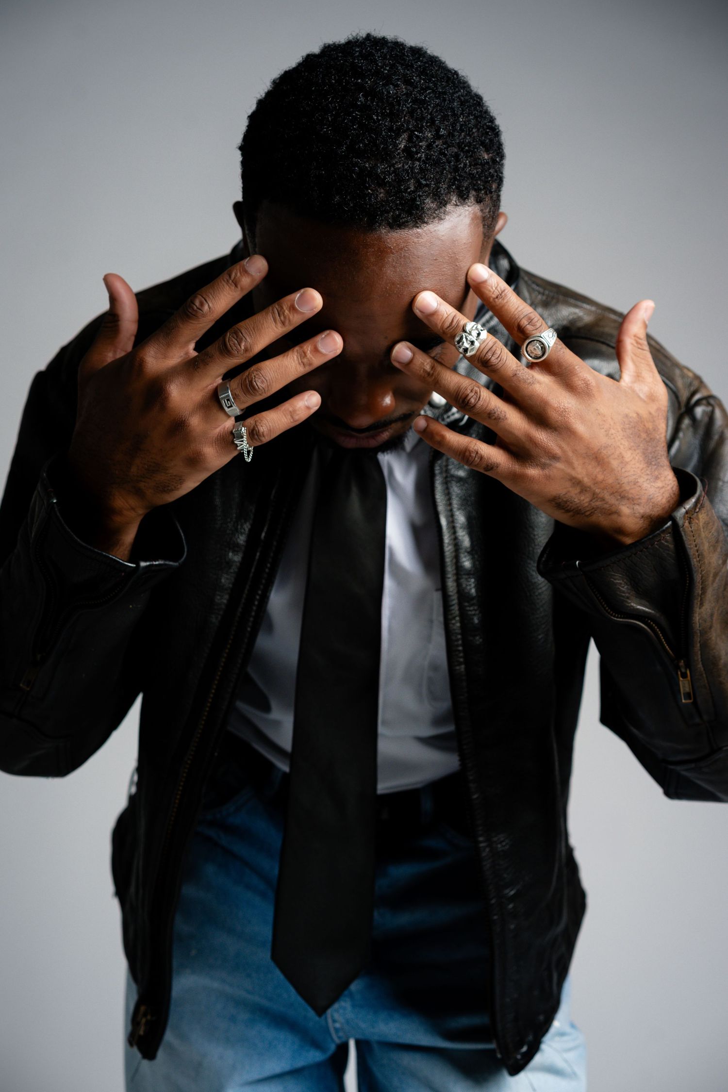 Man in leather jacket and black tie covers face with ring-adorned hands in dramatic pose.