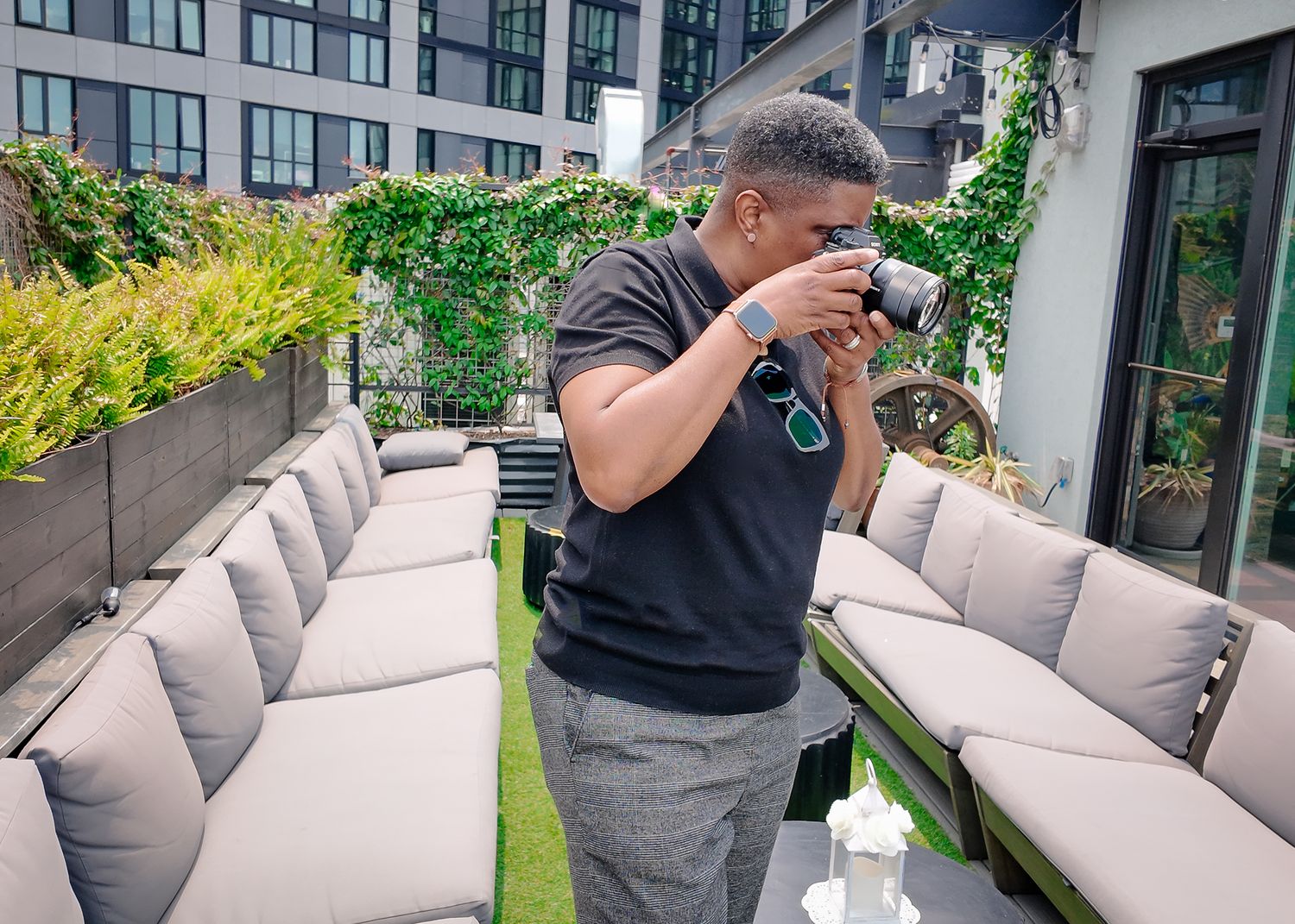 Man in black t-shirt photographs with camera on rooftop patio with lush greenery backdrop.