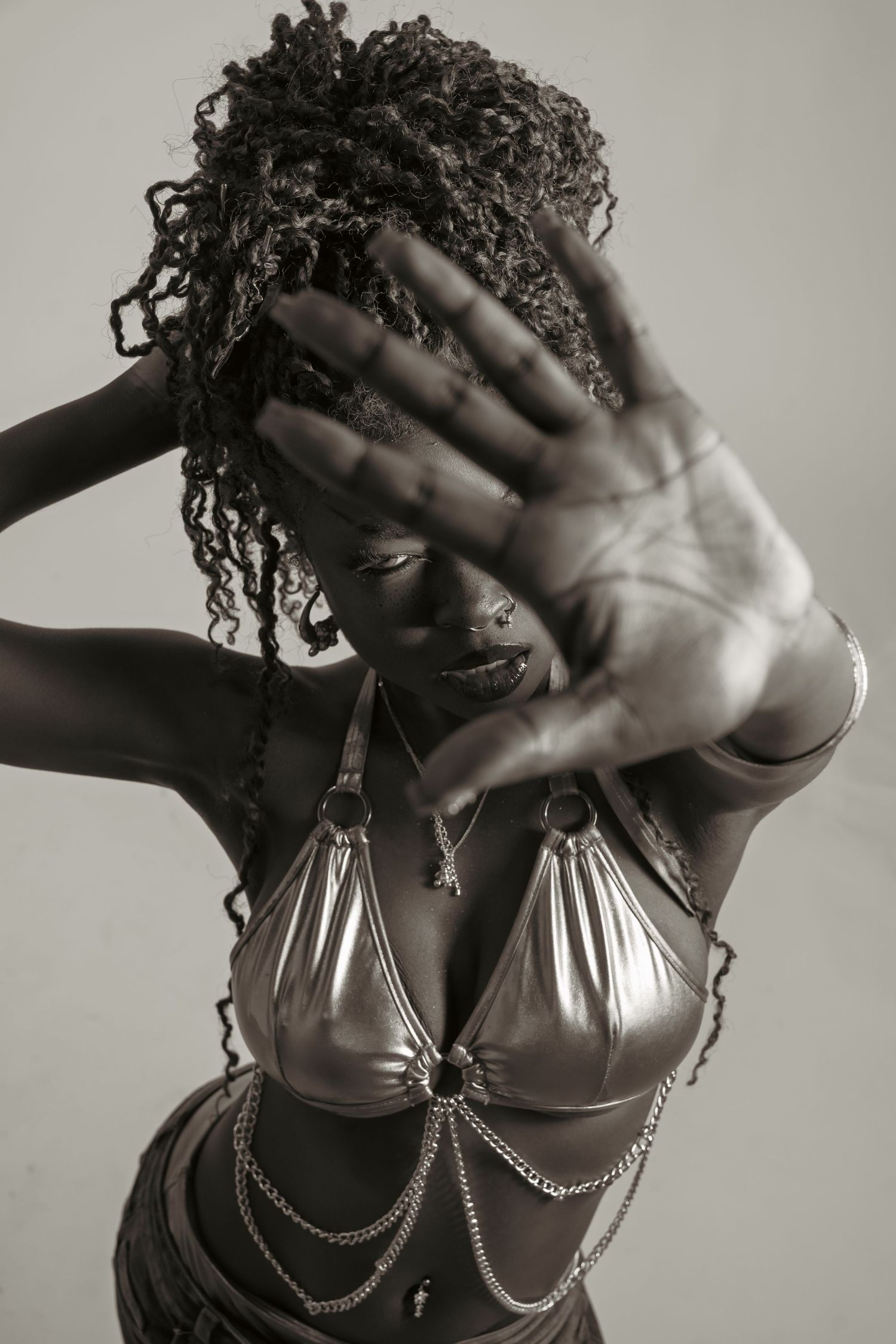 Black and white photo of a woman in silver bikini top with chains, raising hand toward camera with curly hair.