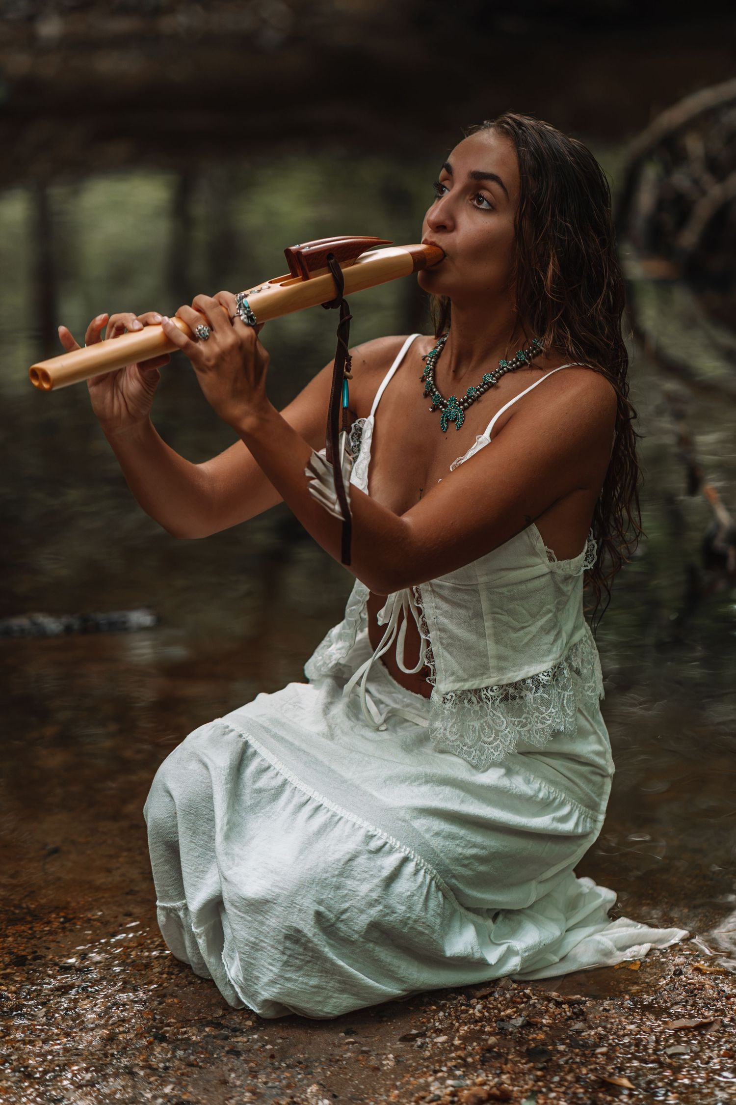 Woman in white dress playing a wooden flute while kneeling in a shallow stream in a forest setting.