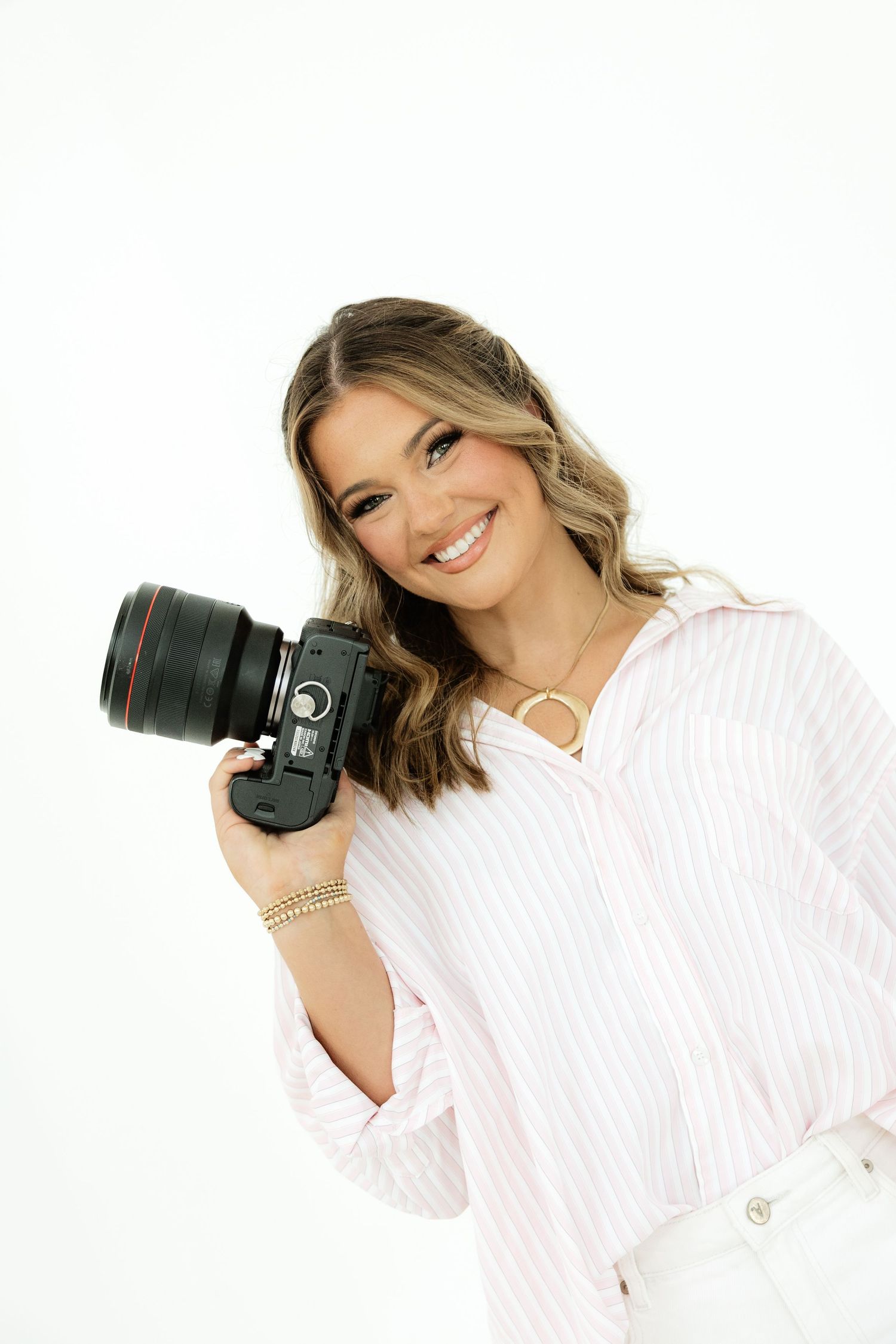 Smiling woman holding a DSLR camera, wearing a pink striped shirt, with long wavy hair on a white background.