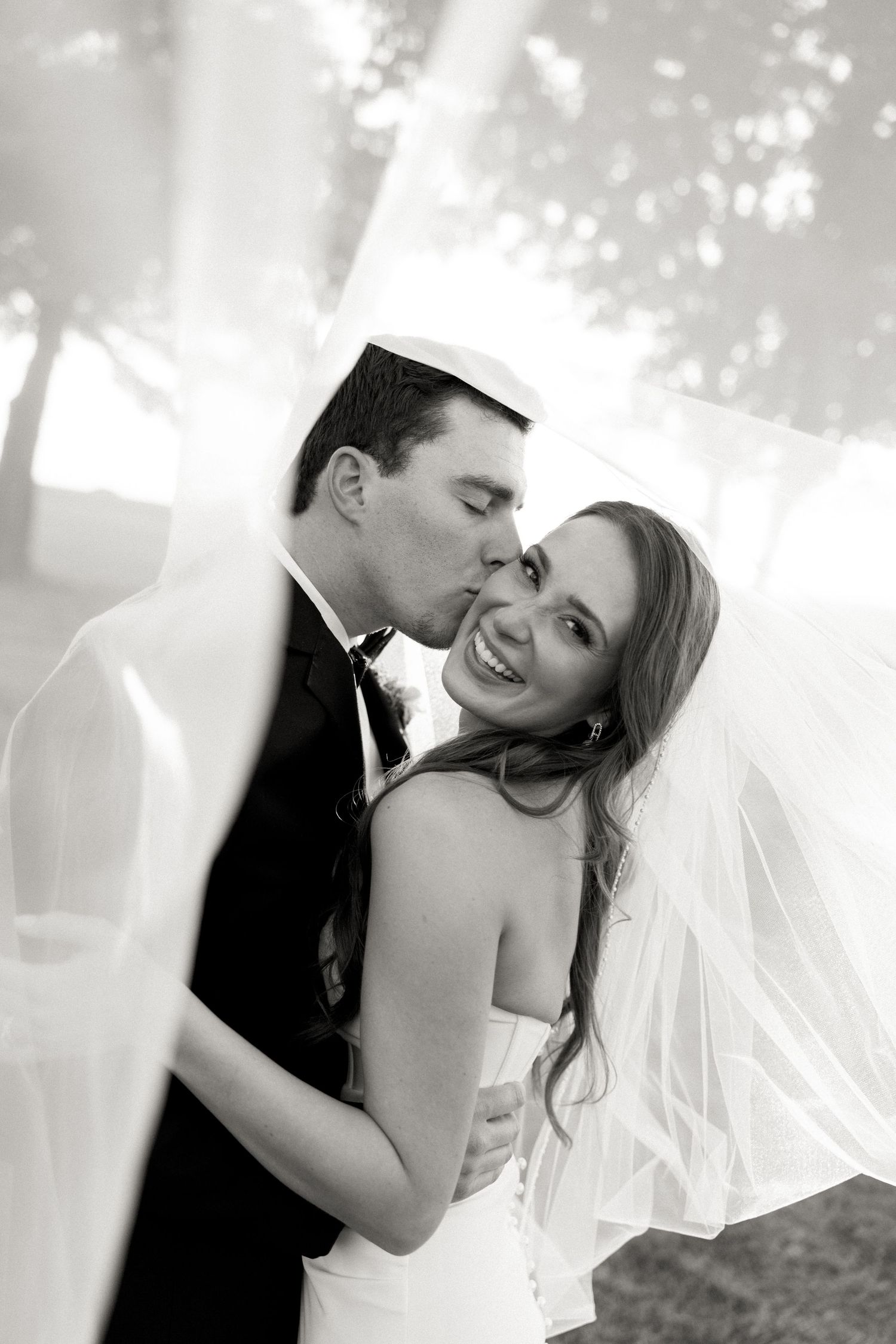 Black and white photo of groom kissing smiling bride on cheek with veil flowing around them.