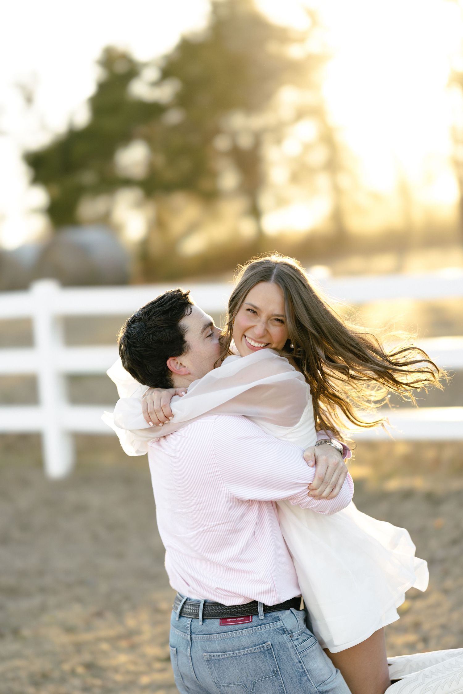 Joyful couple embracing at golden hour near a white fence on a farm.