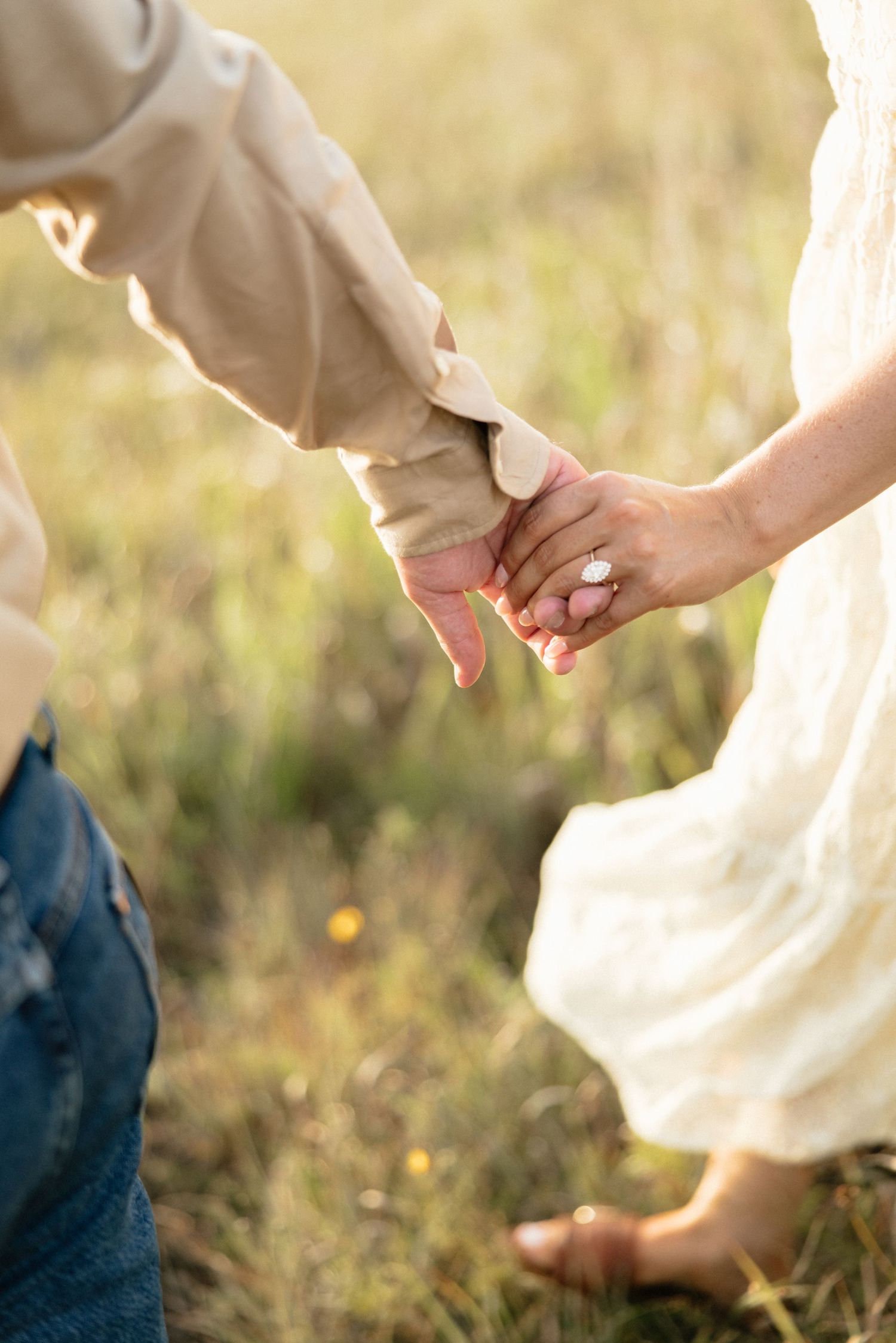 Close-up of couple holding hands in a sunny field, woman wearing white dress with bare feet.
