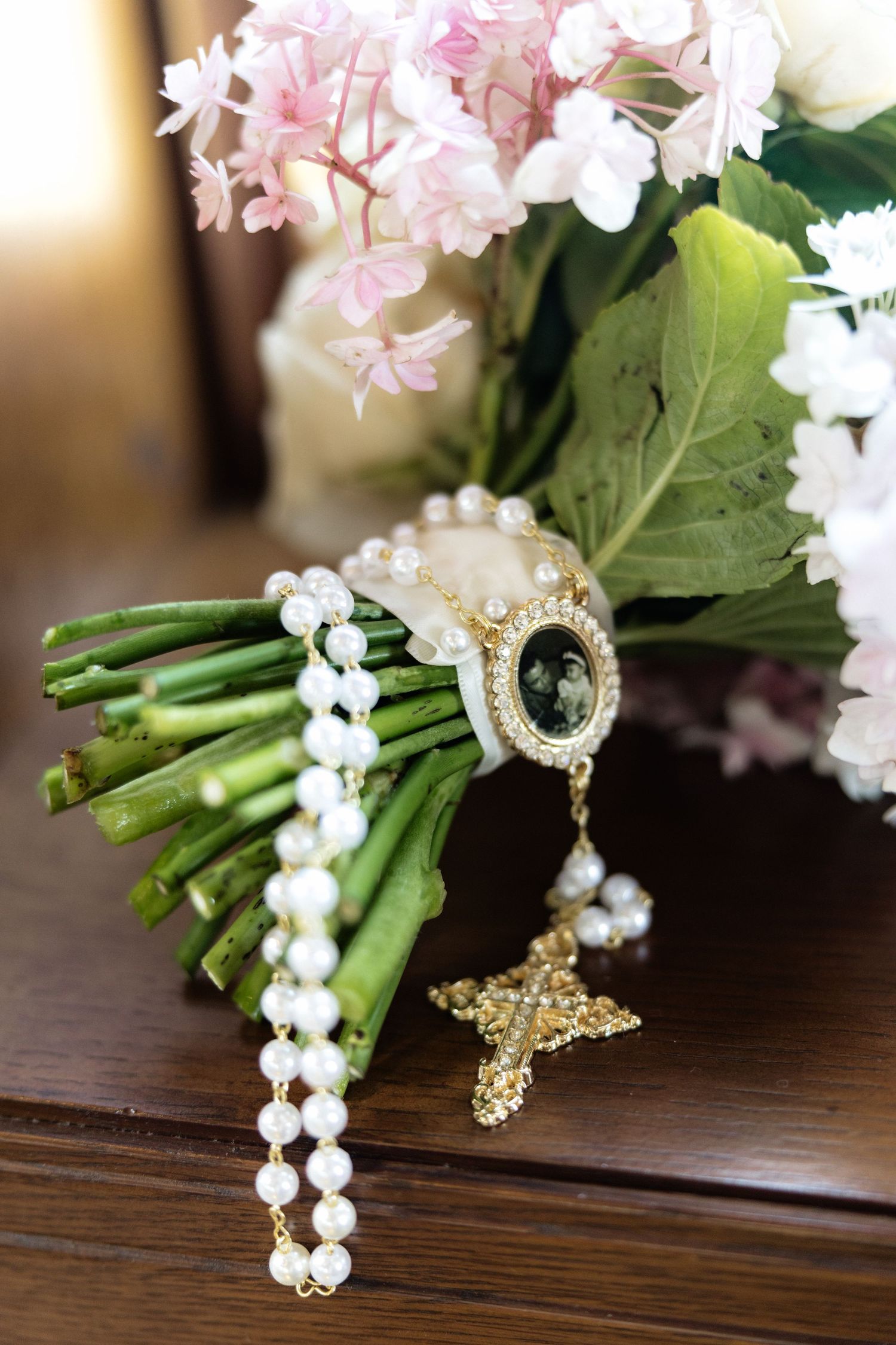 Bridal bouquet with pink hydrangeas and white roses adorned with pearl rosary and vintage photo charm on wooden table.