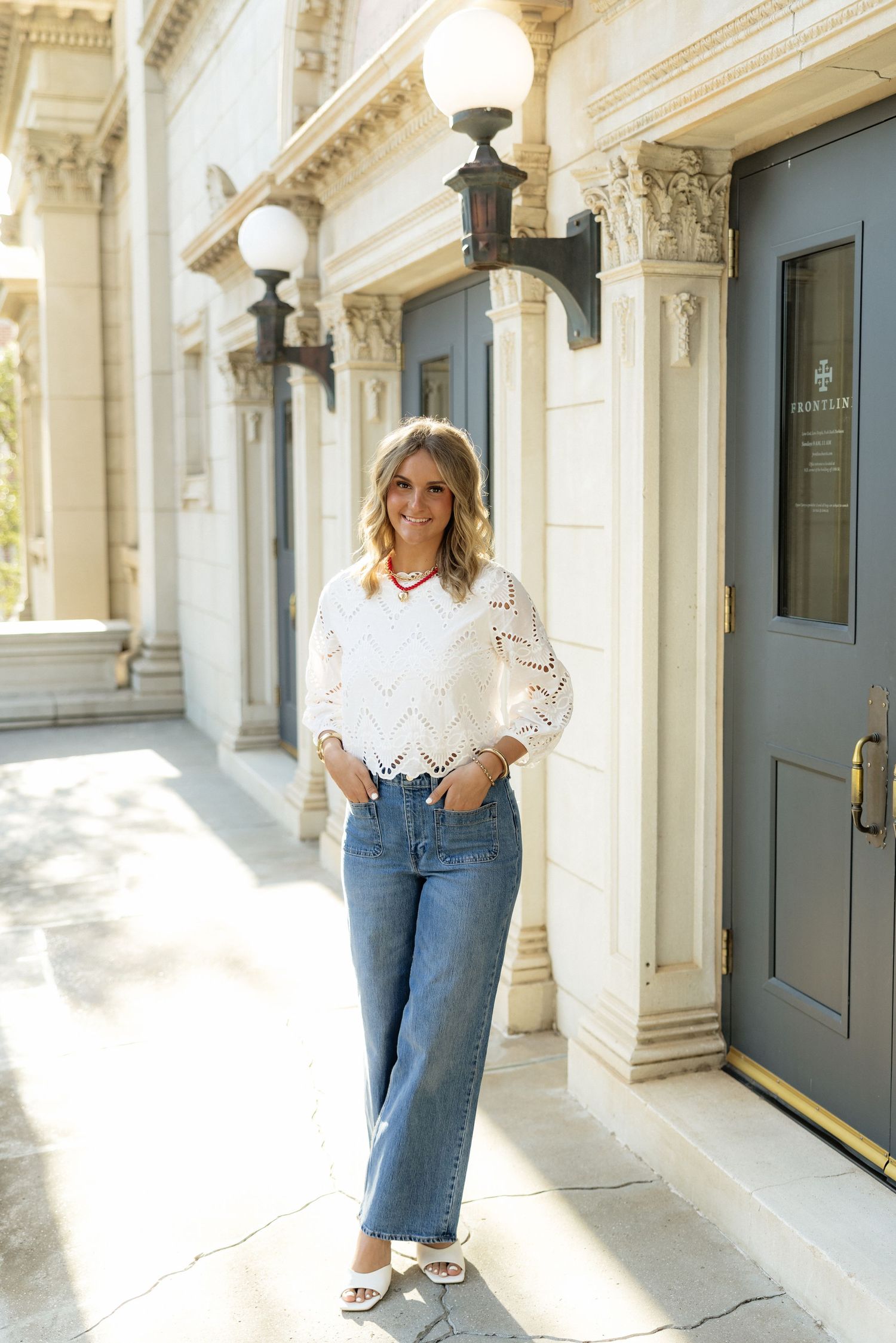 Woman in white lace top and blue jeans posing outside a classic building with ornate columns and a vintage lamp post.