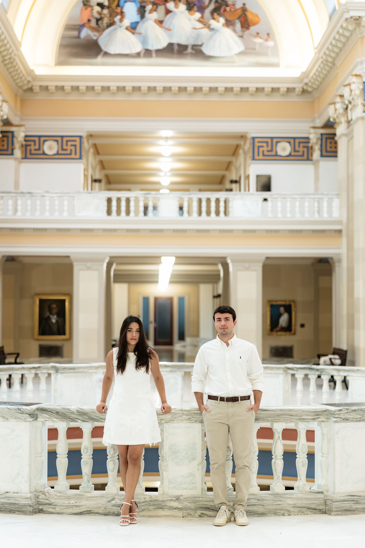 Couple in white attire posing in elegant marble hall with ornate ceiling mural and classical architecture.