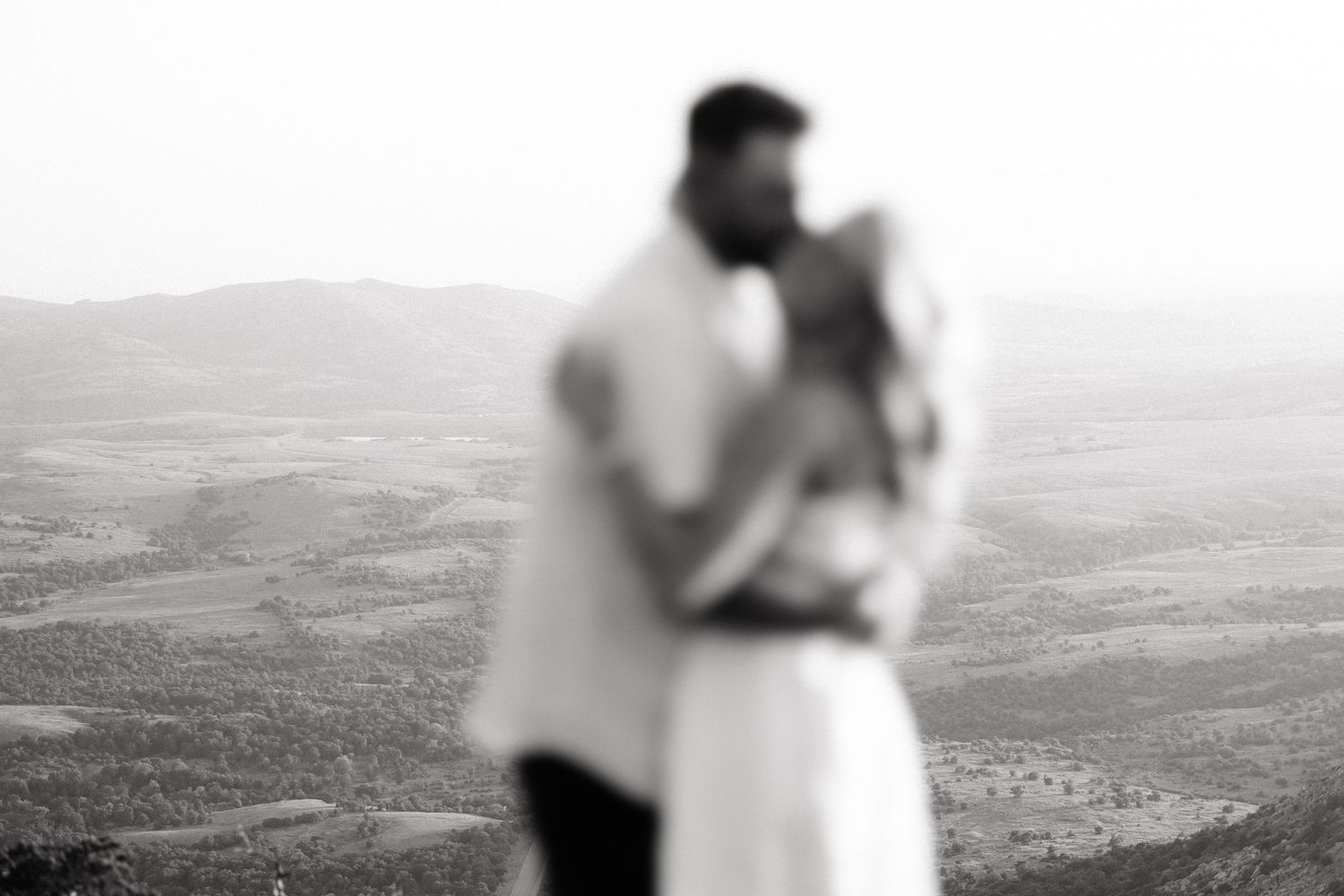 Blurred couple embracing on a hilltop with a sweeping black and white landscape view in the background.