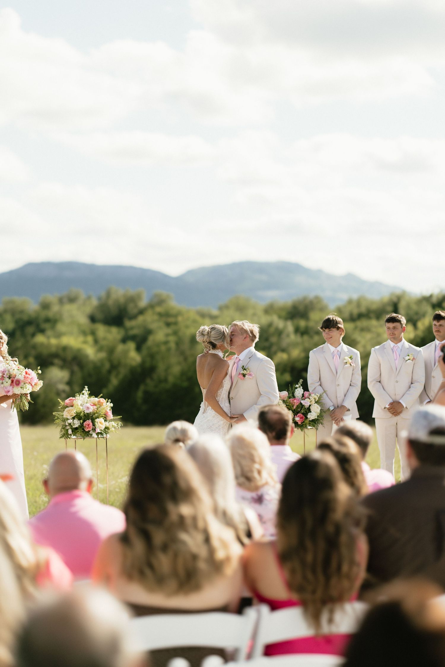Bride and groom share first kiss at outdoor wedding ceremony with mountain views and floral arrangements.