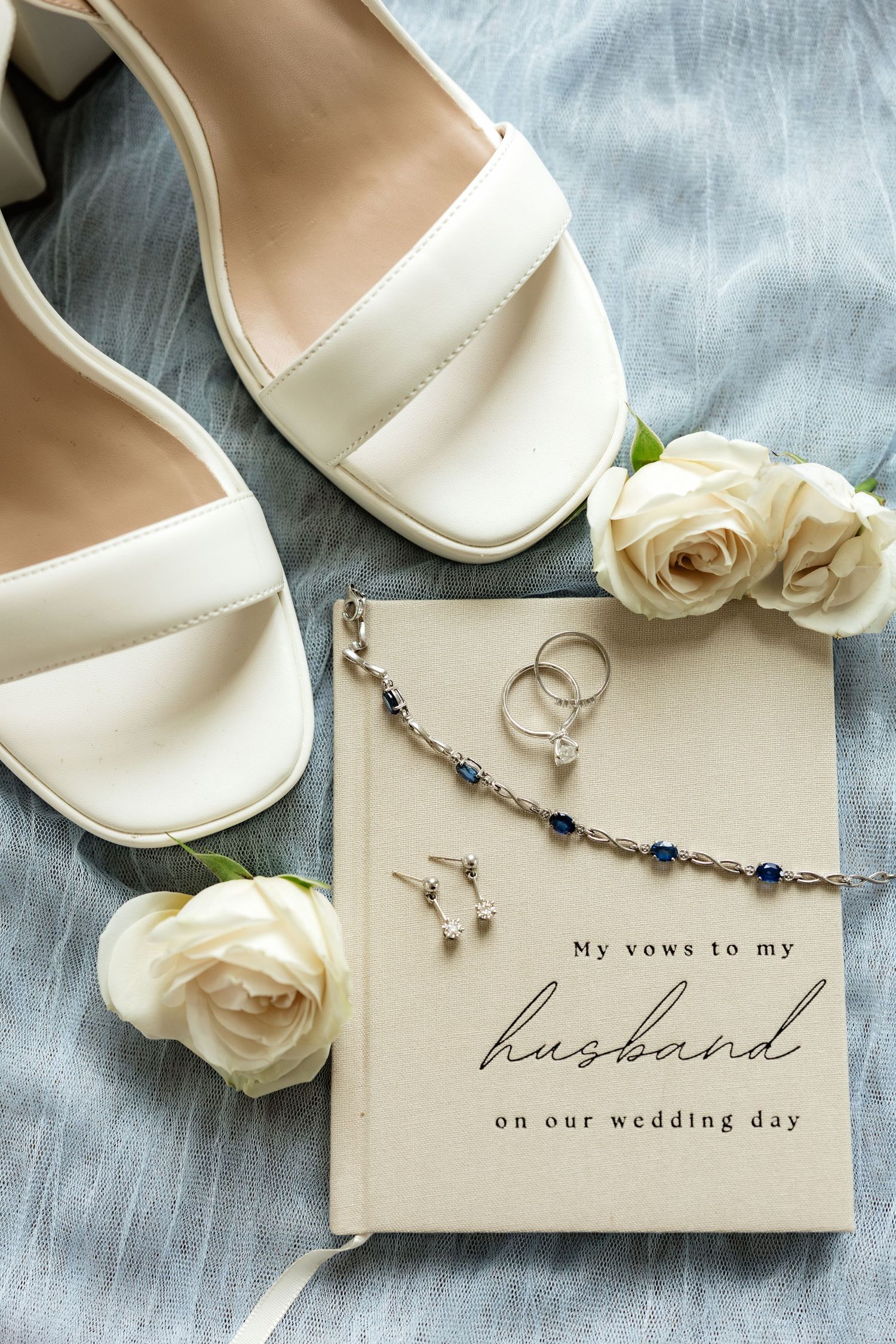 White bridal heels, roses, vow book, and sapphire jewelry arranged on blue linen for a wedding day flat lay.