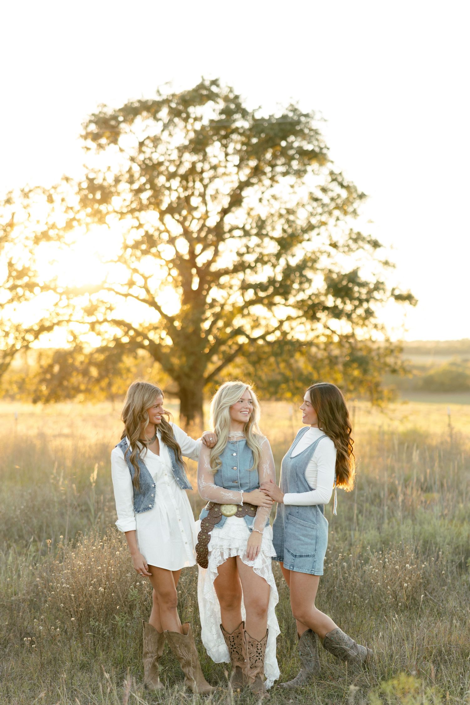 Three women in boho western outfits posing together in golden field at sunset with large oak tree behind them.