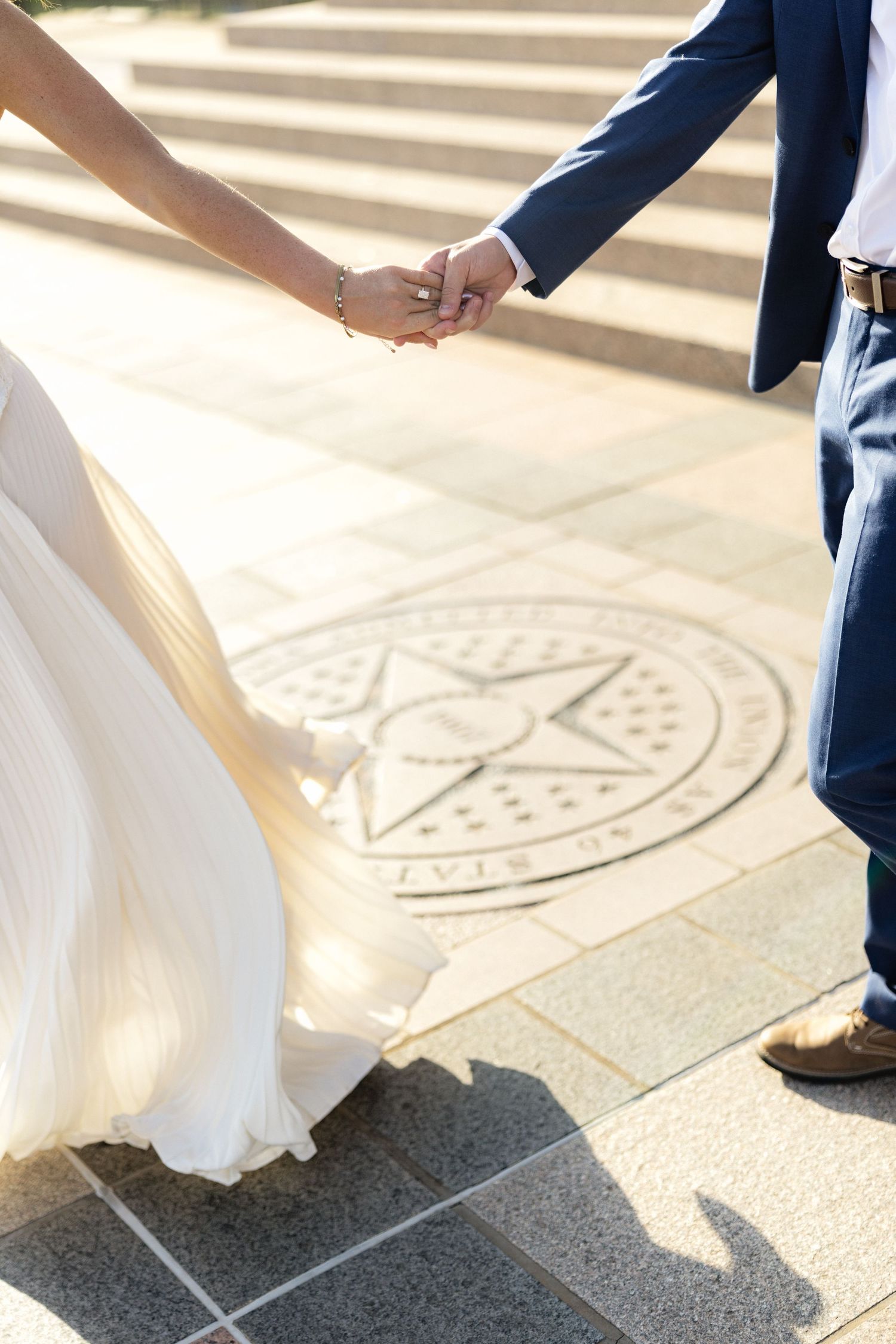 Bride and groom holding hands in wedding attire near a Texas State University seal on stone steps.