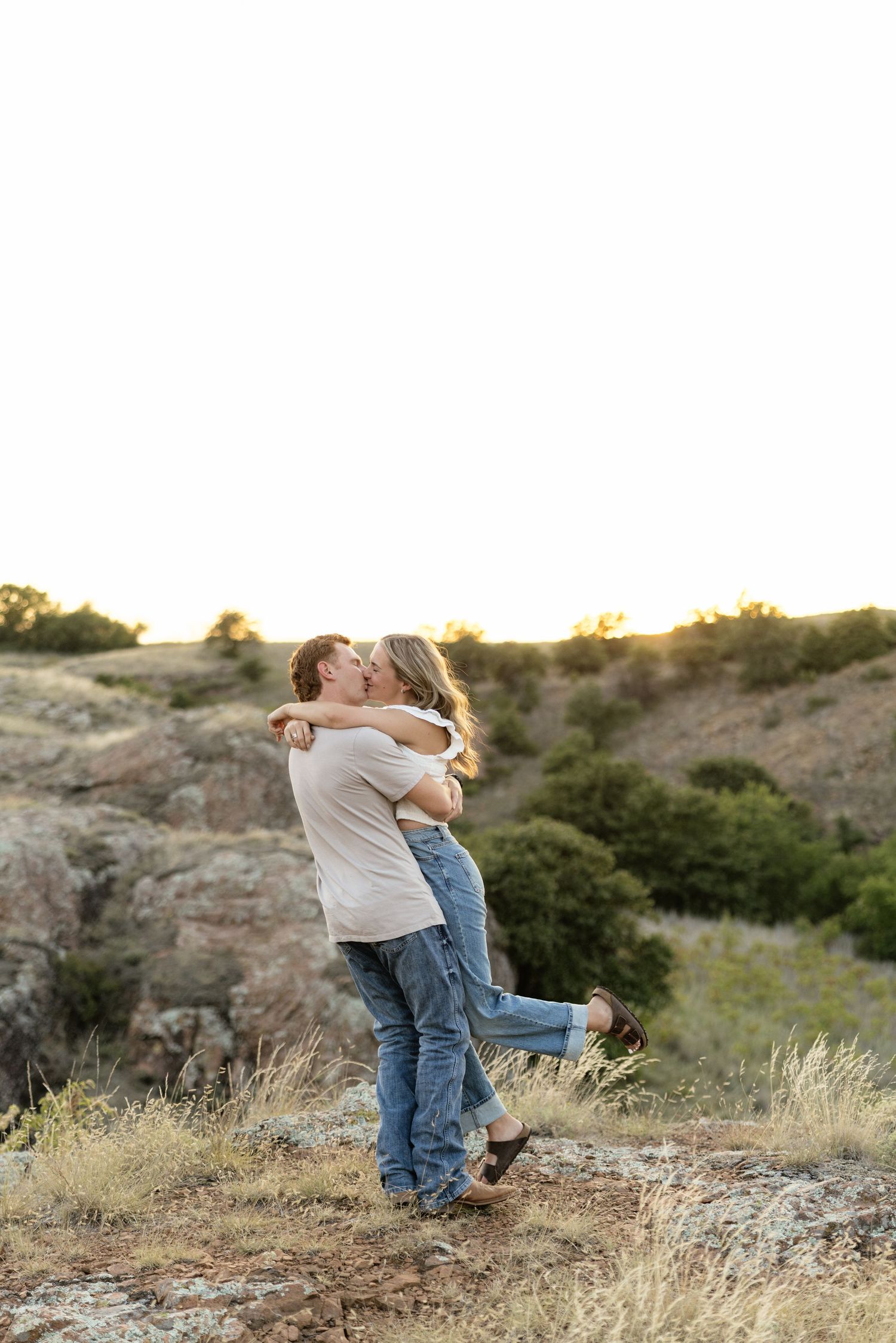 Couple sharing a romantic kiss at golden hour on a rocky overlook with rolling green hills in the background.