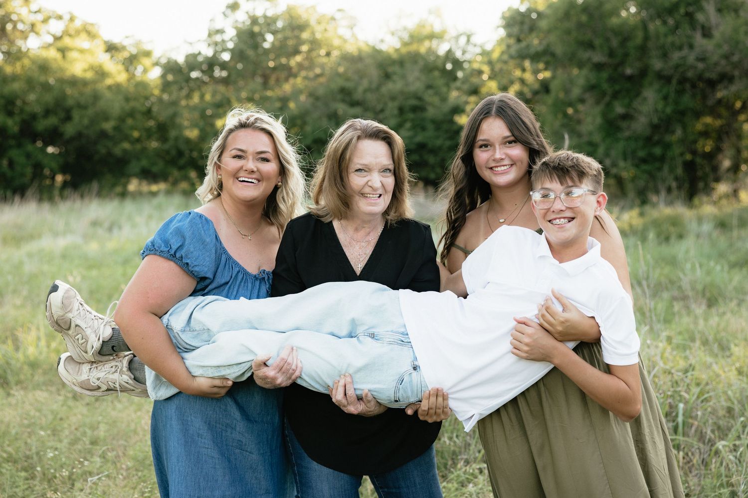 Four people smile outdoors in a field, holding a person wrapped in white, surrounded by lush green trees.