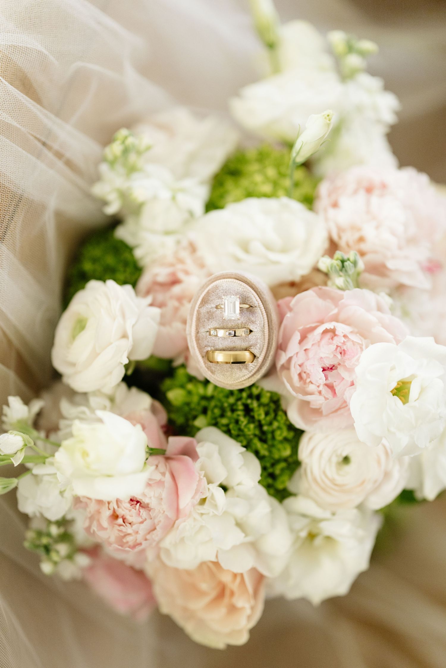 Wedding rings in a velvet box resting on a bridal bouquet of pink peonies and white ranunculus.